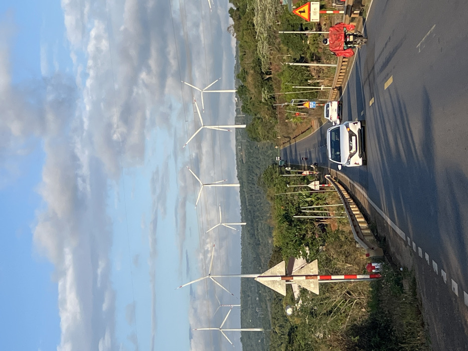 The image depicts a two-lane asphalt road traversing a rural or semi-rural landscape in Ea Ksô, Vietnam, under a sky with scattered white and grey clouds. The road, featuring a yellow double solid line and white dashed shoulder lines, is slightly elevated with metal guardrails on both sides and appears to ascend into the distance.

In the background, at least eight large, white, three-bladed wind turbines are positioned on rolling green hills, some partially obscured by the terrain or clouds. Overhead, multiple power lines stretch across the sky.

On the road, a white four-door sedan travels towards the viewer in the center-left lane. Behind it, another white sedan moves away from the viewer. On the right shoulder, a person on a motorcycle, draped in a prominent red covering over their back, travels away from the viewer. Further down the road, additional vehicles, including a motorcycle and a truck, are visible.

The roadside on the left features a red and white striped pole with two triangular signs, along with dense green vegetation and a partially visible white building. On the right, a yellow triangular traffic sign with a black arrow indicating a left turn and "400m" below it is present, alongside utility poles and more green foliage. Long shadows stretch across the road from the left, indicating late afternoon or early morning sunlight.