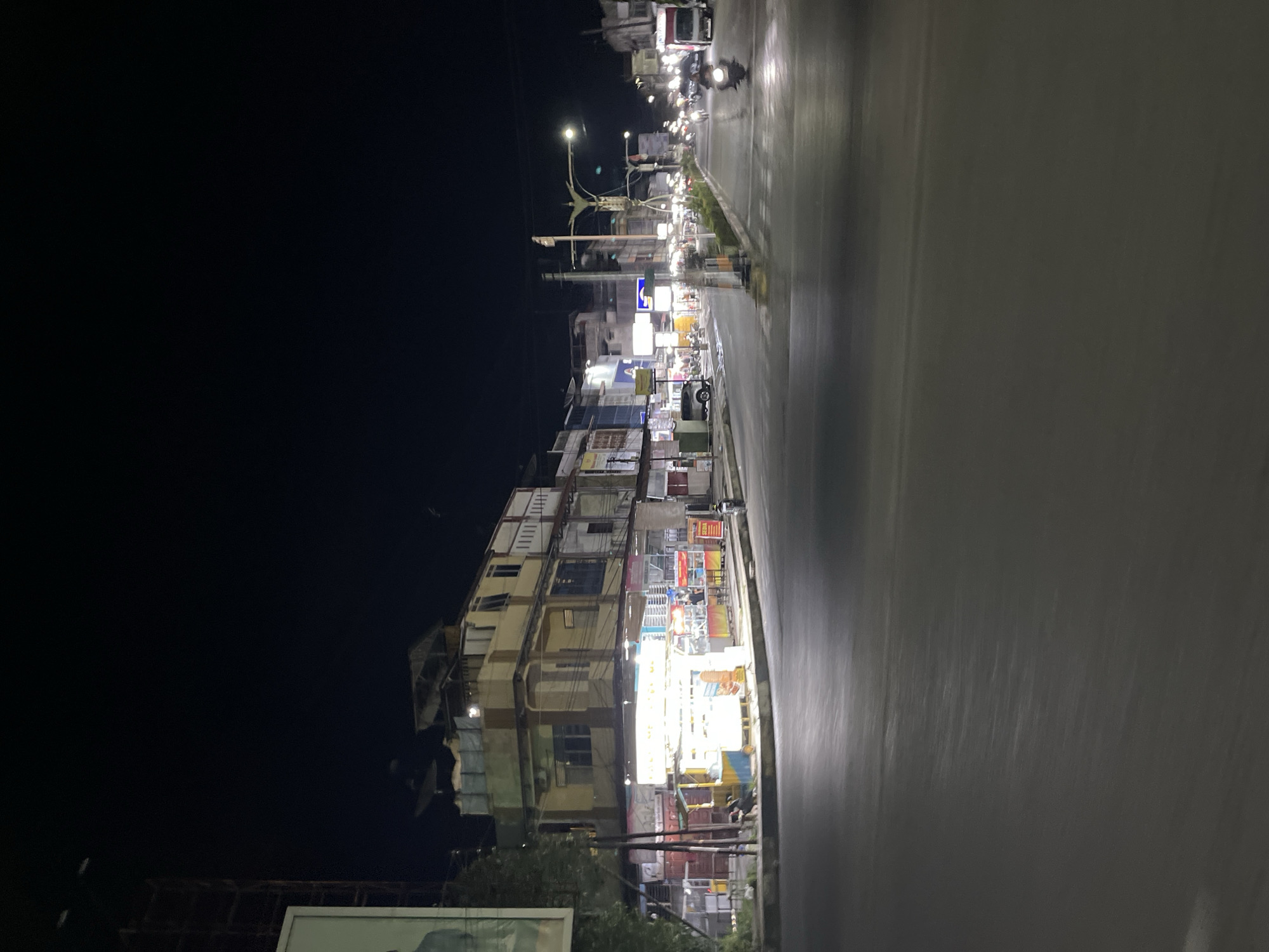 This is a night-time, medium-angle, vertical shot of a street in Langsa, Indonesia. The image captures a commercial area with a multi-story building lining the left side of the street. The building appears to be a mix of residential and commercial spaces, with illuminated storefronts at ground level. Several food stalls are visible, some with bright lights, indicating they are open for business. A few individuals are present, some possibly staff at the stalls, and others potentially customers. Vehicles are present, including a motorcycle in the foreground on the right, and other cars and trucks further down the street, some of which are in motion, suggested by motion blur. The street itself is paved, and streetlights illuminate the scene. The overall atmosphere suggests a bustling urban environment, even at night.