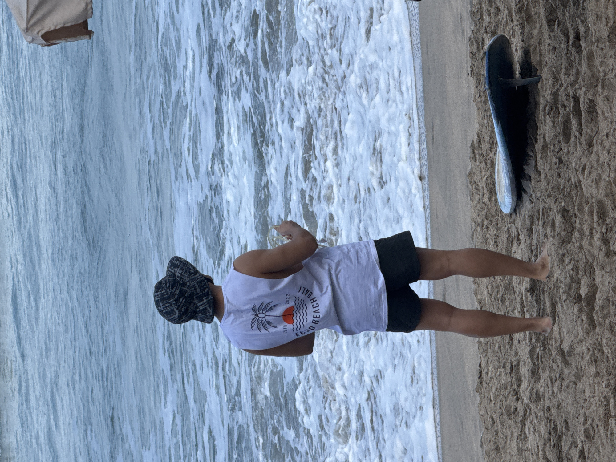 A person wearing a bucket hat and a white tank top stands on the sandy shore of Kuta Beach in Kuta, Indonesia, facing the ocean. The tank top has a graphic of a palm tree with a sunset over waves and text that reads "ESTD 1977 2022 ECHO BEACH BALI." The person is holding a clear glass in their right hand. To the right of the person, a blue surfboard is propped up on the sand. The ocean has white foam from waves. In the background, a beige beach umbrella is partially visible.