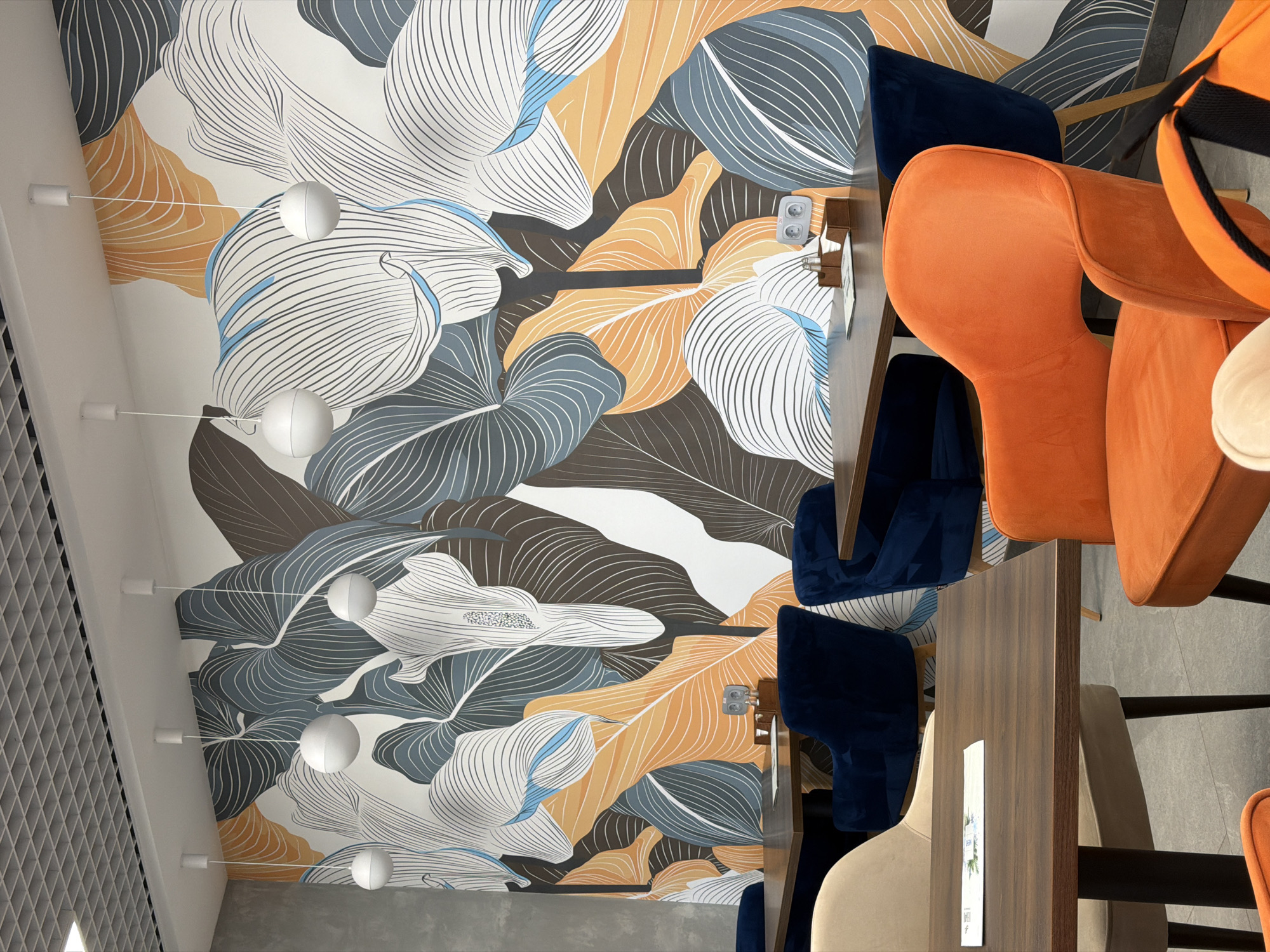 The image depicts the interior of a restaurant, featuring tables and chairs with a decorative wall design. The tables are dark brown wood, set with minimalist menus. Chairs surrounding the tables include orange, dark blue, and beige velvet seating. The walls are decorated with a stylized botanical motif composed of large leaves in shades of white, blue, brown, and orange. Several spherical pendant lights are suspended from the ceiling. The location is Chaikovsky, Russia. No people are visible in the frame.