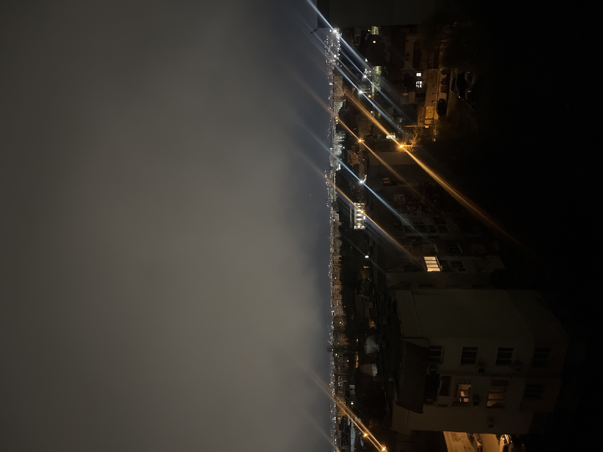 The image is a nighttime cityscape, likely taken from a high vantage point. The left side of the image is dominated by heavy fog or low-lying clouds, obscuring details below. The right side of the image shows urban development with numerous buildings and streetlights illuminating the scene. The perspective is unusual, as the image is rotated 90 degrees. The location is plausibly Sofia, Bulgaria, based on architectural styles, urban density, and street lighting characteristics. No people are visible.