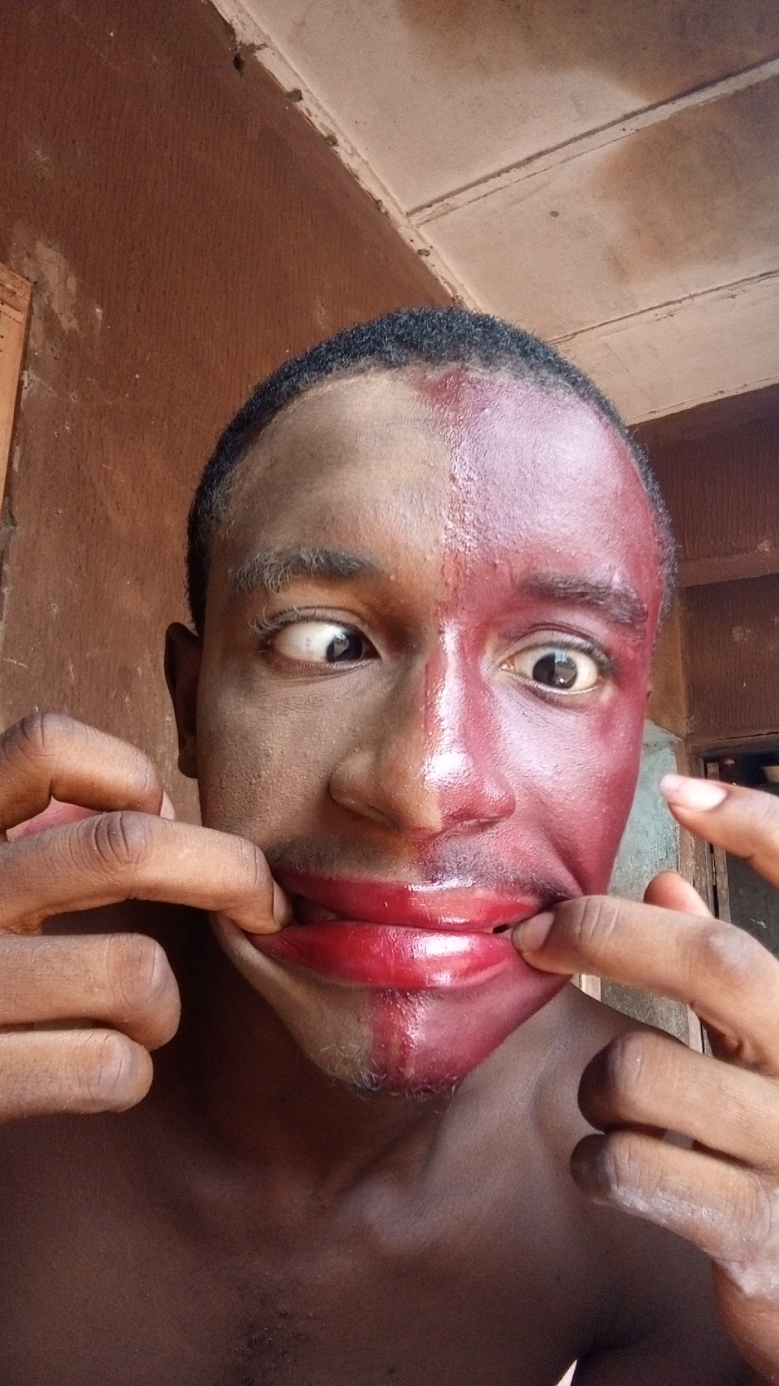 A close-up shot of a dark-skinned man with face paint, likely in Onitsha, Nigeria. The face paint is split down the middle, one side a reddish-brown hue and the other a muted brown. His eyes are slightly crossed. His fingers are pulling the corners of his mouth outwards, creating a stretched smile. The background is a wall with signs of age and wear.