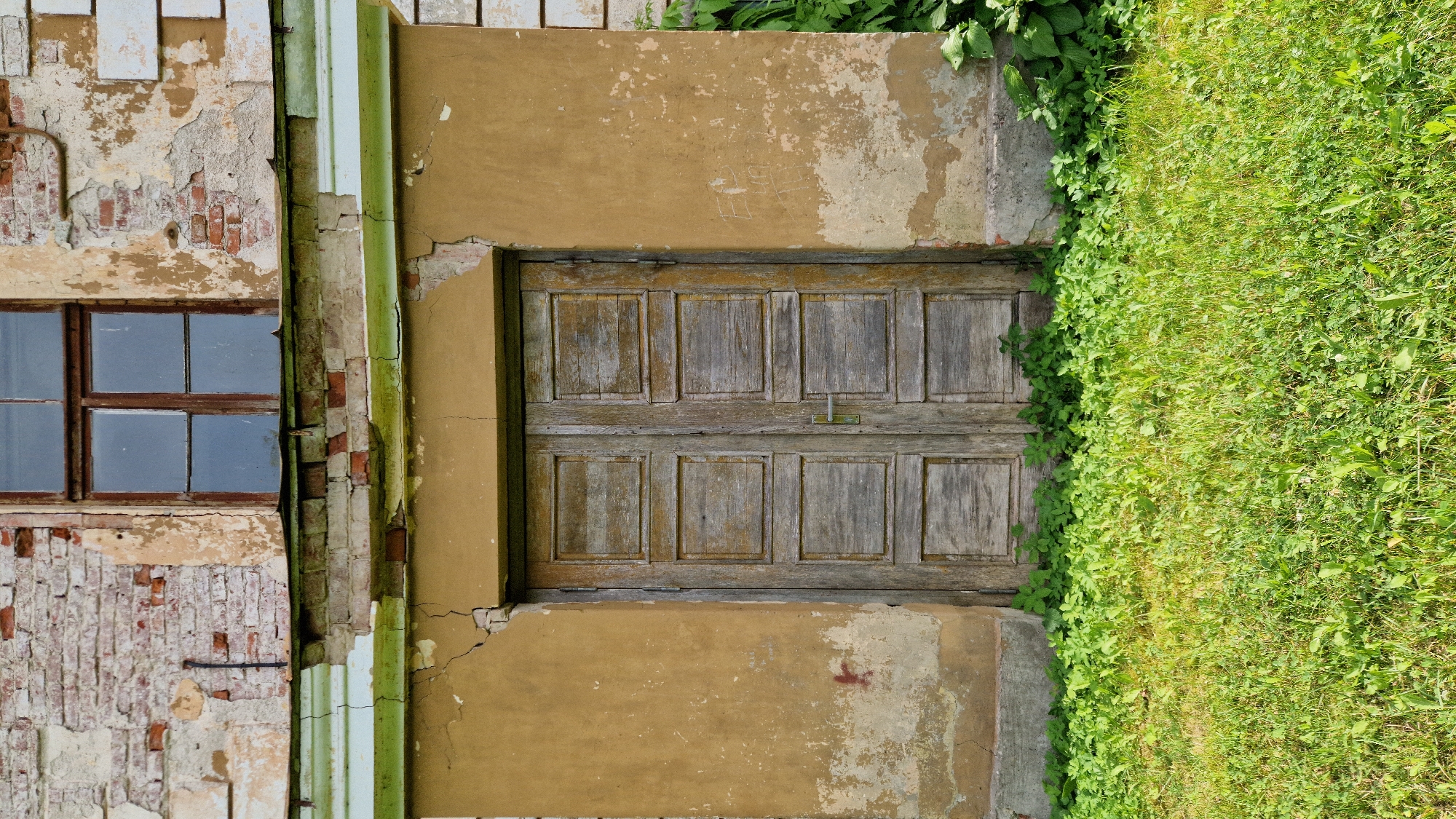 The image shows an exterior view of an old, weathered building with a distressed facade in Kazdanga, Latvia. A double wooden door, aged and showing signs of wear, is centrally located in a mustard-yellow, stucco-covered section of the building. Above the door, a window with a dark frame and multiple panes is visible within a section of exposed brickwork and crumbling plaster. The lower portion of the building features overgrown green grass and foliage, suggesting neglect or a natural landscape reclaiming the structure. The overall impression is one of decay and disuse, characteristic of historical sites that have been left to the elements. There are no people present in the image.