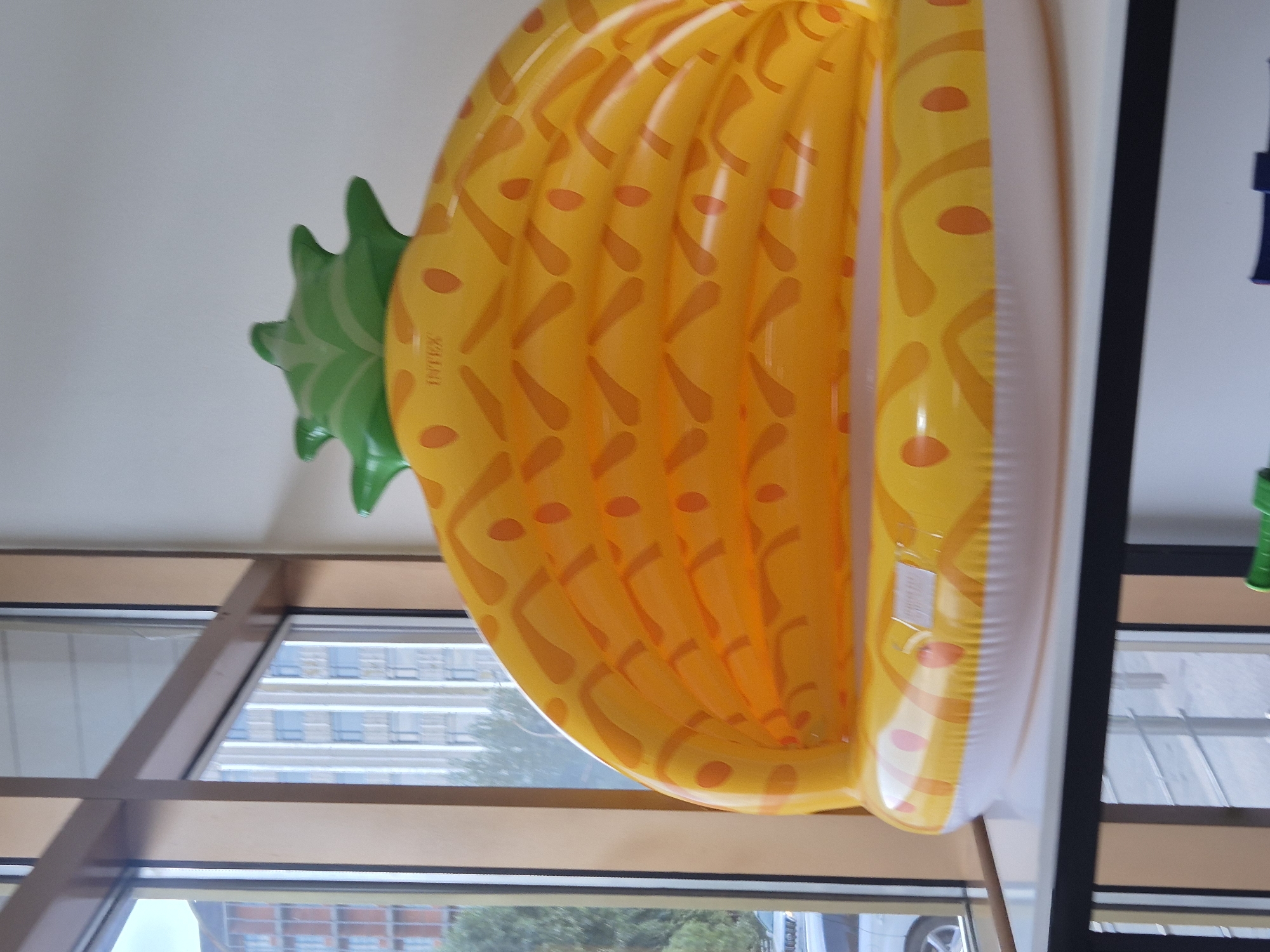 An inflatable pineapple-shaped pool, predominantly yellow with orange diamond patterns and green leaves, is positioned indoors on a white surface. The pool has a white inflatable rim with some pink accents. Behind the pool, a window reveals a cityscape with buildings and trees. The scene appears to be a retail or display area. The location is not specified in the image or its context.