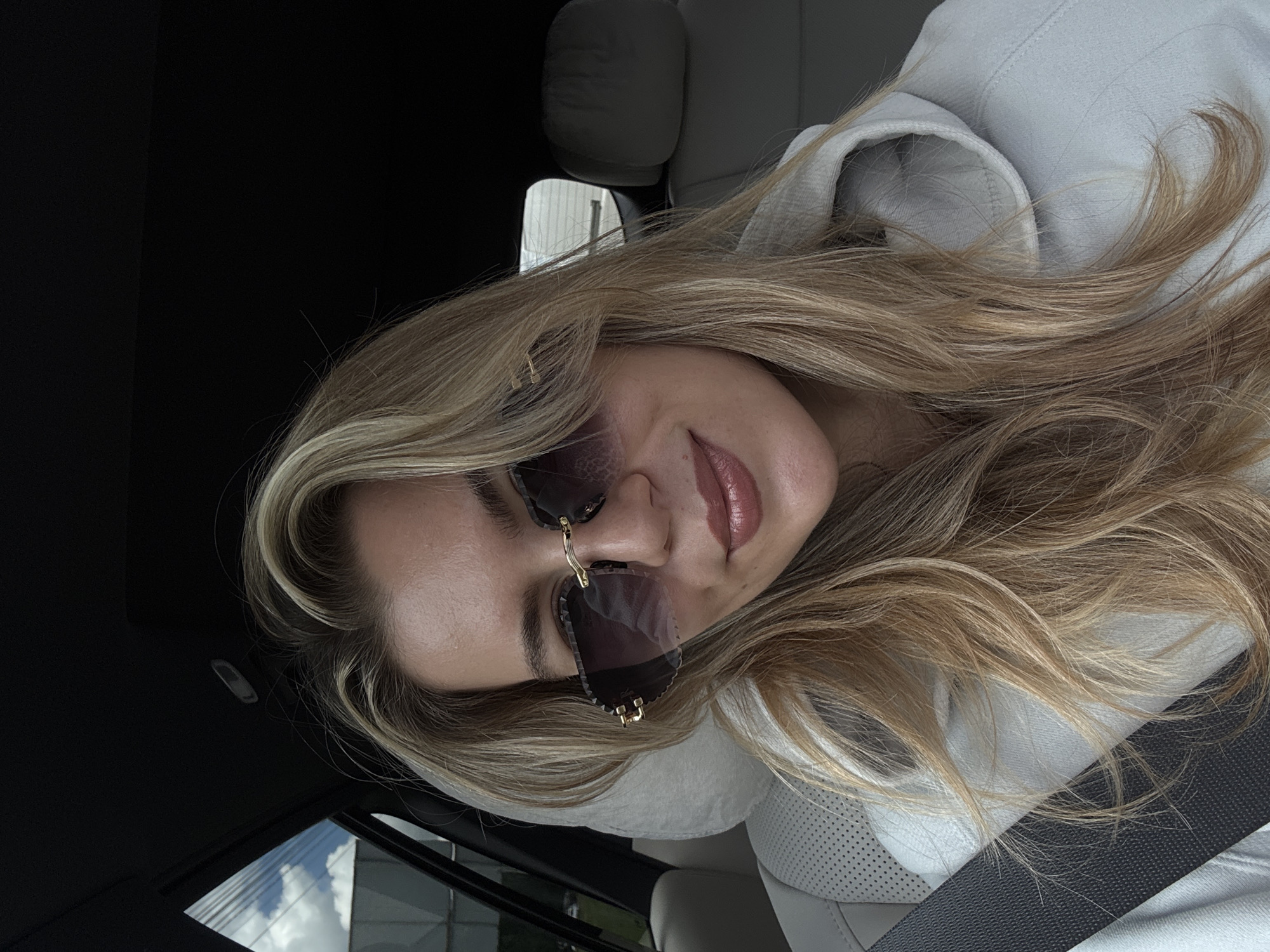 A young woman is captured in a close-up portrait, likely a selfie, inside what appears to be a car. She has long, wavy blonde and light brown hair that cascades around her face and shoulders. Her head is tilted slightly, and she looks towards the camera with a gentle, closed-mouth smile.
She is wearing dark, possibly rimless, sunglasses featuring a distinctive gold-colored bridge and accents on the temples. Her lips are adorned with a natural, glossy pink or nude lipstick. She is dressed in a light-colored hooded sweatshirt, visible around her neck and shoulders, and a dark seatbelt is fastened across her chest.
The background elements confirm her location in a vehicle. A light grey, perforated headrest is visible behind her, and the car's ceiling is dark. Through the windows, bright daylight illuminates the scene, reflecting a sky with white clouds and parts of a modern building with horizontal architectural lines. The overall impression is one of a casual moment during a car journey on a bright day. No visible text is present in the image.