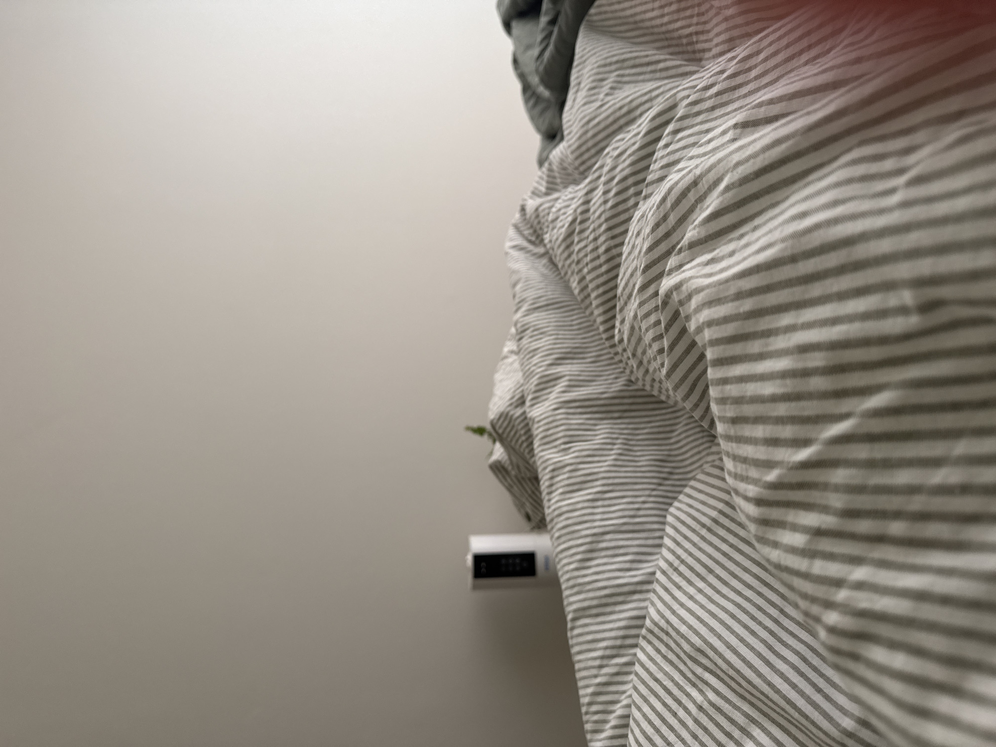The image shows a bedroom with a striped duvet cover dominating the right side of the frame. A white digital clock is mounted on the gray wall to the left of the bed. The rest of the left side of the image is a plain gray wall. The location, Люк, Russia, is not discernible from the visual elements.
