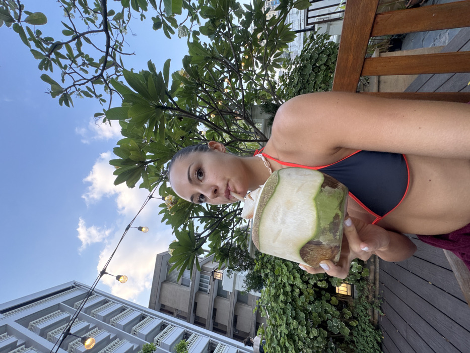 A young adult female is depicted in a medium-shot photograph on an outdoor terrace in Bangkok, Thailand. She has light skin, dark hair pulled back, and is wearing a black bikini top with red trim and a shell choker necklace. She holds a large, light-green coconut with a white straw to her mouth, drinking from it while looking directly at the camera. Her left arm and upper torso are visible, with a dark red towel or garment draped below her waist. The immediate environment includes dark wooden decking and a wooden bench or railing to the right. String lights with exposed bulbs are suspended overhead. The background features a partly cloudy blue sky, dense green tropical foliage, and trees bearing light-colored flowers. To the left, a multi-story white high-rise building with multiple balconies is visible, with another building behind it. A dark lantern-style fixture is affixed to a wall within the greenery. A partial figure is discernible in the distant background near the base of the white building.