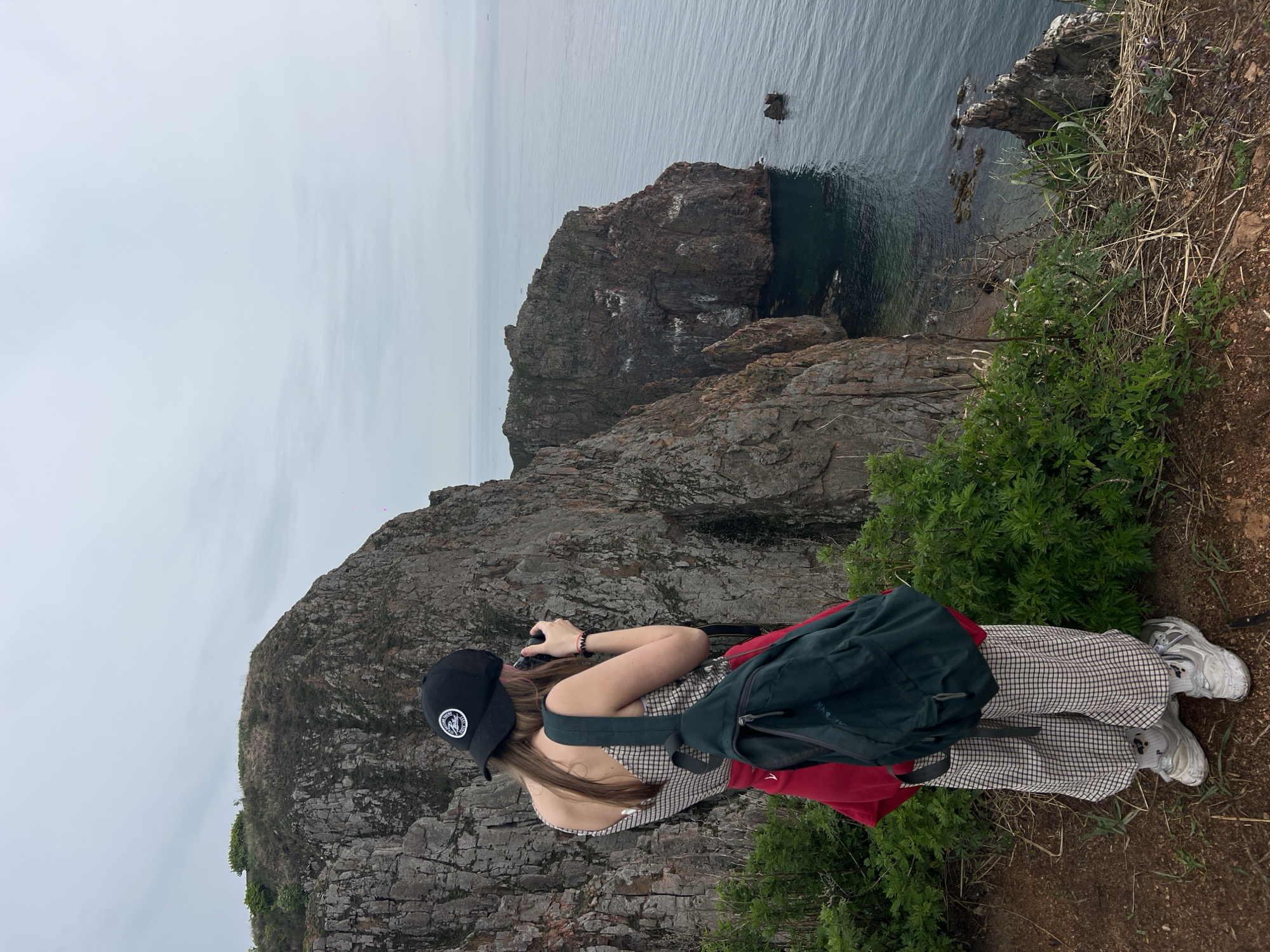 A person stands on a cliff's edge, presumably in Russky Island, Russia. The person is looking through a camera. They are wearing a black baseball cap, a checkered dress, a green backpack, and white sneakers. A red jacket is visible beneath the backpack. The rocky cliff falls down to the ocean, with an additional large rock formation in the water. The sky is overcast. Vegetation grows along the cliff's edge.