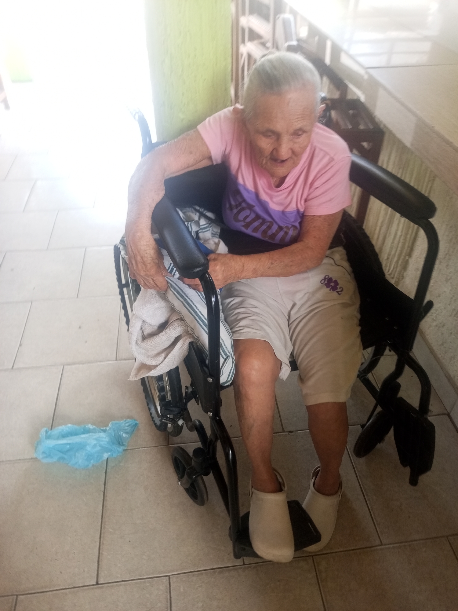 An elderly woman with grey hair is seated in a black wheelchair on a tiled floor. She is wearing a pink t-shirt with purple lettering and light-colored shorts. A light-colored towel is draped over her lap and the armrest of the wheelchair. To the left of the wheelchair, a crumpled light blue plastic bag is on the floor. The environment appears to be indoors, with a bright light source illuminating the scene from the left. The background consists of a green wall and a wooden railing. The overall impression is that of a person in a wheelchair, possibly at home or in a care facility, in Juan Griego, Venezuela.