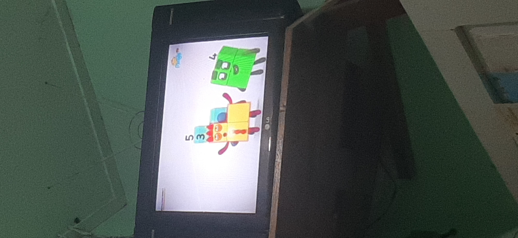 A television screen displays animated characters from the show "Numberblocks." On the left, a character composed of yellow and red blocks stacked to represent numbers 5 and 3, with blue blocks at the top, has red arms and legs. To the right, a green block character representing the number 4 stands with a smiling face. The television screen has the LG logo at the bottom. The television is placed on a stand, which is below a dark surface. The background is a textured green wall.