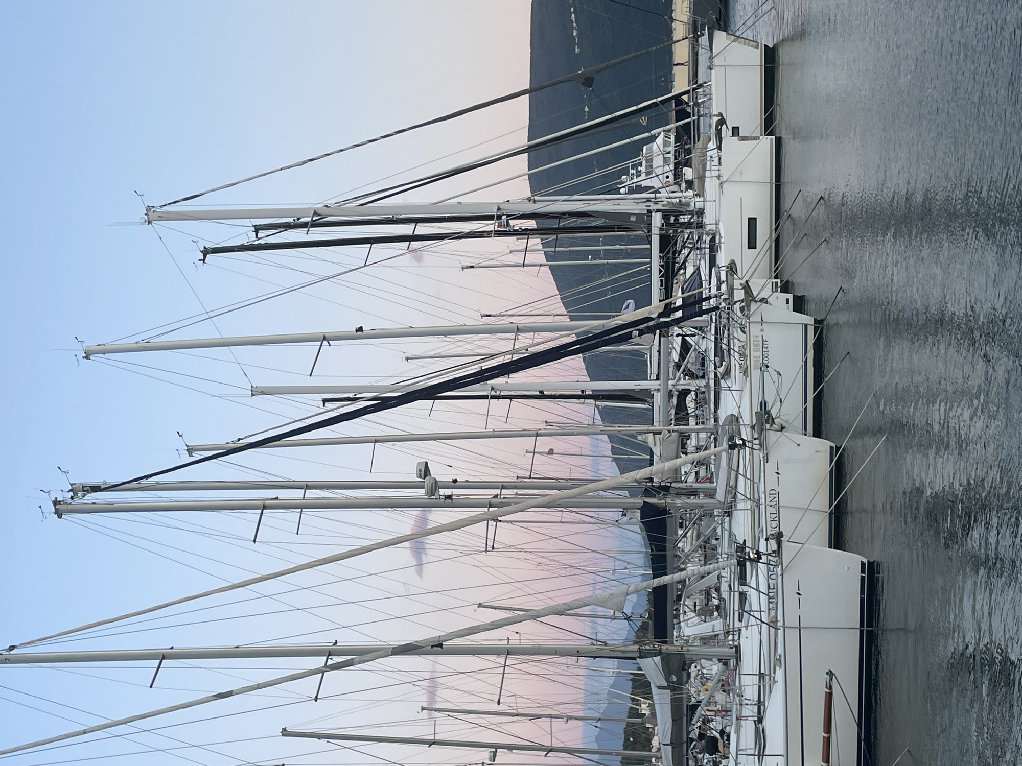 The image shows a close-up view of sailboats moored in a marina. The masts of the sailboats dominate the scene, creating a dense network of lines against a sky transitioning from blue to pink hues, possibly at dawn or dusk. Some boats are labeled "RICKLAND". The marina is located in Meljine, Montenegro. The water reflects the boats and masts. In the background, a large, dark ship is partially visible. No people are discernible in the image.