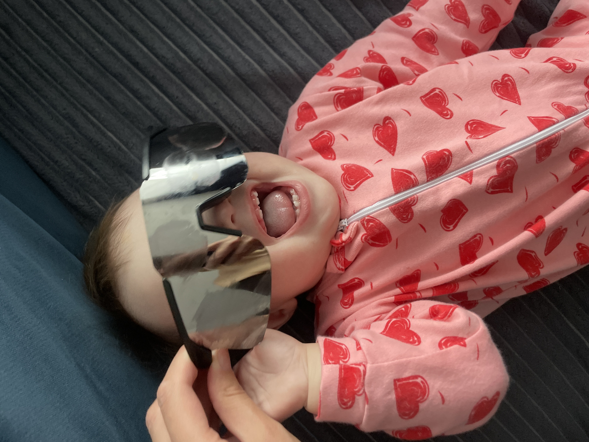 An overhead shot captures a baby lying on its back, viewed from an angled perspective. The baby is wearing a light pink onesie patterned with numerous red hearts and features a white zipper down the front. Its mouth is wide open, revealing its tongue and some small teeth. A pair of large, highly reflective, dark-framed glasses or a mask is held over the baby's upper face, obscuring its eyes and nose. An adult's hand is visible holding the glasses in place, while the baby's tiny hand is also near the reflective surface.
The setting appears to be indoors in Podshivalovo, Russia. The baby is resting on a dark gray, ribbed fabric surface, likely a couch or bed. A section of smooth, dark blue fabric is visible in the background on the left side of the frame. The scene suggests a playful moment between the baby and an adult. No discernible text is visible in the image.