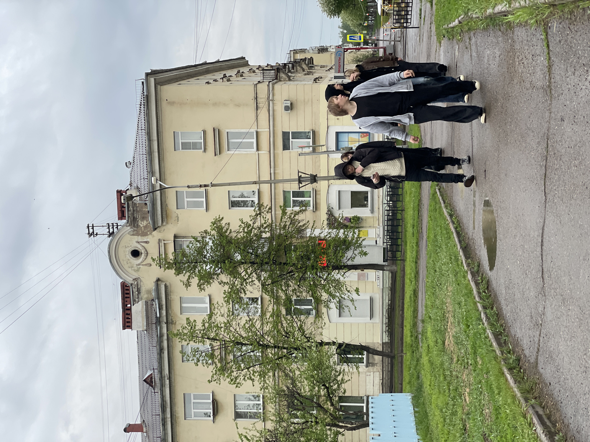 The image shows a group of four people walking on a sidewalk in Zheleznogorsk, Russia. They are walking in the same direction on the right side of the image. The person closest to the camera is wearing a black jacket, black pants, and brown shoes. The person in the middle is wearing a gray sweatshirt and black pants. The building behind them is a multi-story structure with a yellow facade. There are several windows and trees obscuring some of the building's features. The sky is overcast. The street has a puddle of water. A street sign is visible in the background.