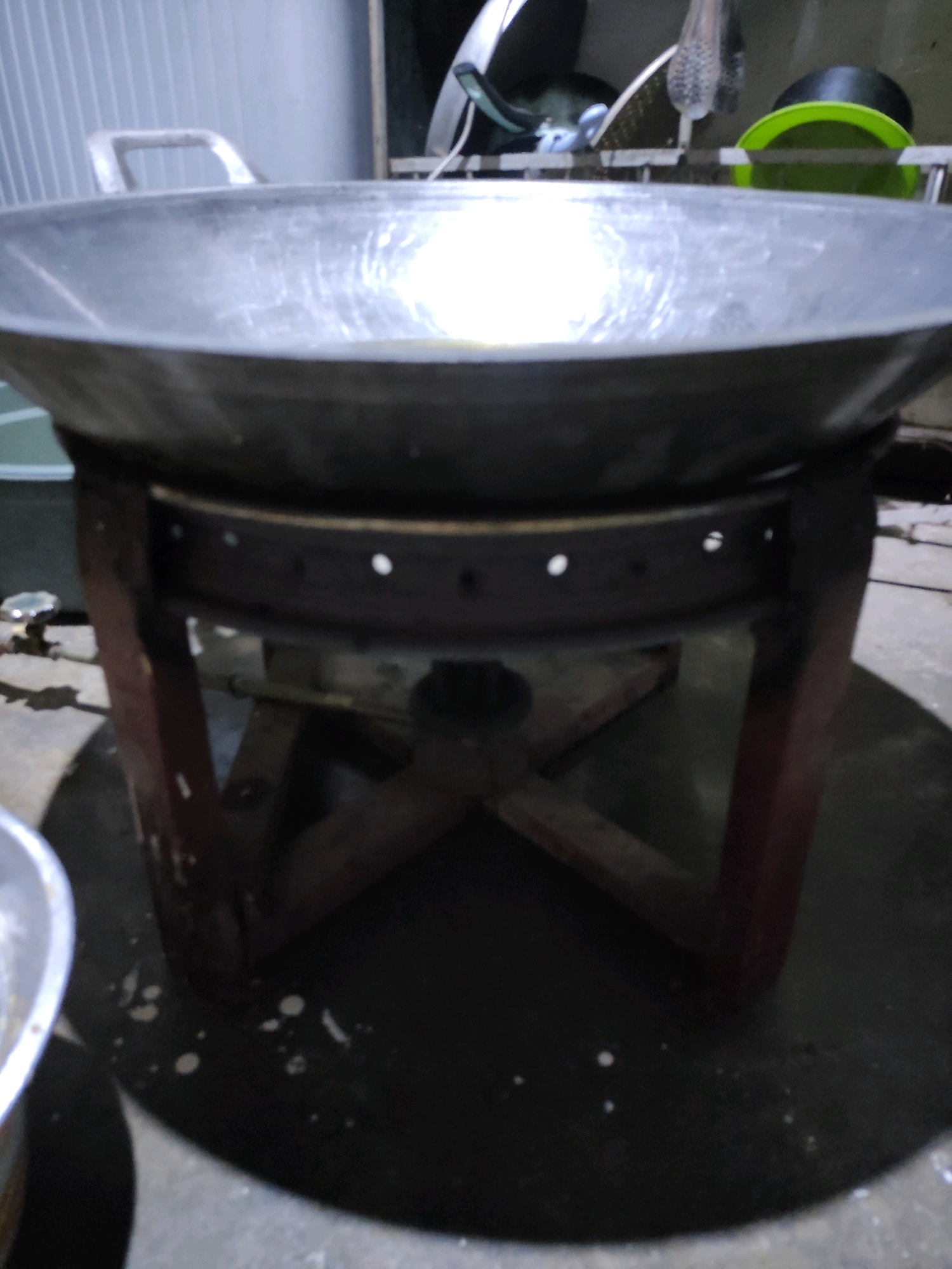 This is a close-up, dimly lit, eye-level, indoor shot of a wok resting on a wooden stand over what appears to be a burner. The wok is made of metal and has two handles. The stand is made of dark wood and has a circular metal rim with small holes around it, supporting the wok. Below the rim, a gas pipe and valve are visible, suggesting the presence of a stove. The background is blurred and appears to be a domestic setting with some kitchen items, including green and black bowls or containers. The floor is a dark, circular surface, possibly a mat or a built-in stove base, with some white specks on it. The overall impression is of a cooking area, likely in a home kitchen in Bulukumba, Indonesia, given the prompt's context.