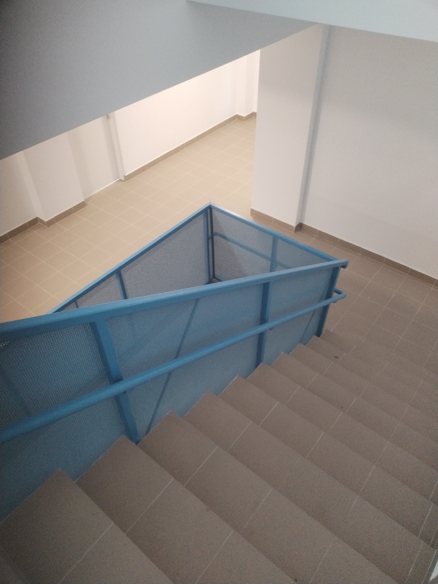 This is a forensic, matter-of-fact description of an interior scene. The image shows a staircase with light brown tiled steps and a light blue metal railing with mesh infill. The railing encloses the stairwell. The walls are painted white, and the floor in the landing area at the top of the stairs is tiled in a light beige color. The ceiling appears to be painted a light gray. The scene is clean and appears to be in good condition. There are no people or animals visible in the image. The lighting is bright and even, suggesting artificial lighting. The architecture suggests a modern, public or commercial building. The location of Bucharest, Romania is not visually apparent from the image itself and would require external context.