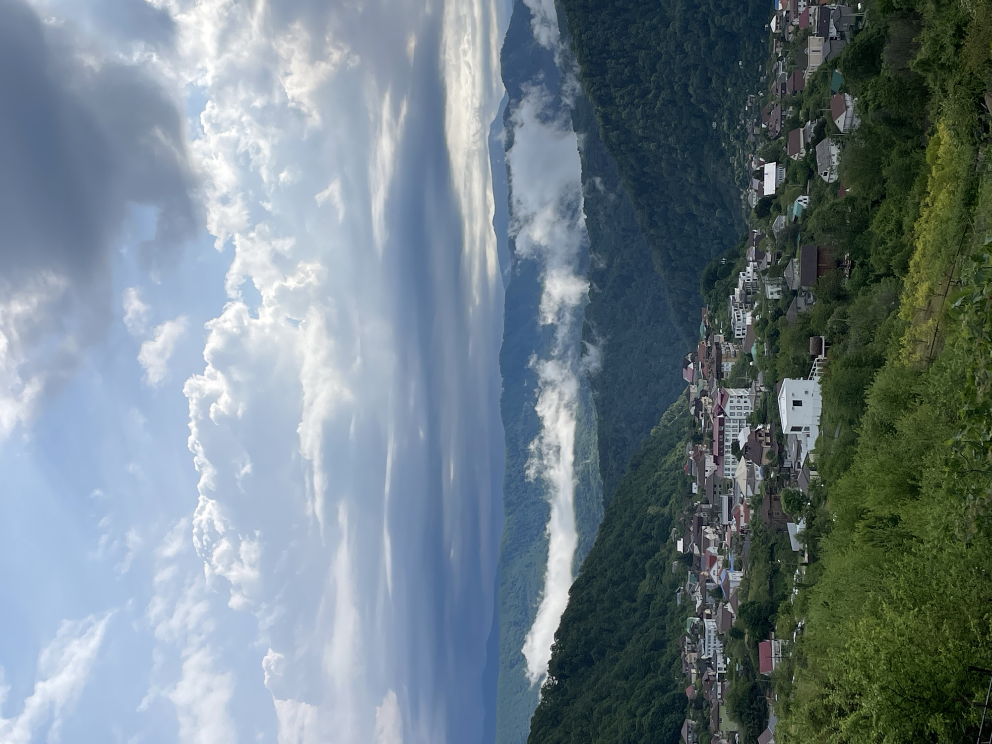 The image shows a mountain village on the slopes of a green, densely forested mountain in the Sochi region of Russia. Buildings, mostly white with red and green roofs, are scattered across the hillside. The sky, split between blue with white, puffy clouds and a darker, ominous grey, suggests an approaching storm. Lower mountains are visible in the background, partially obscured by fog. The viewpoint is elevated, providing a wide view of the scene.