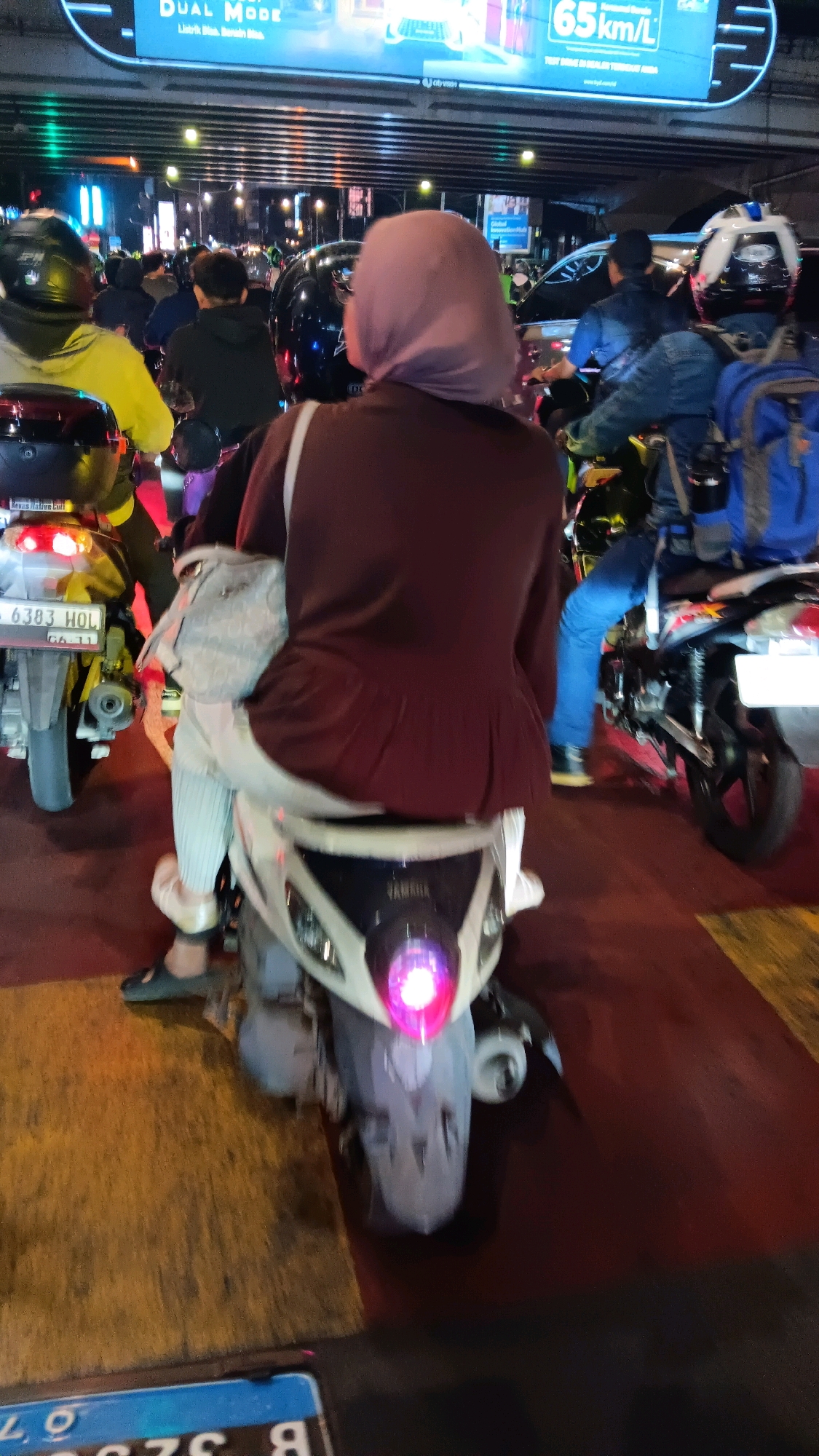 This image depicts a street scene in South Jakarta, Indonesia, likely during nighttime or dusk, characterized by heavy traffic. In the foreground, a woman wearing a light purple hijab and a maroon sweater is riding on the back of a white scooter. She is wearing striped pants and dark slip-on shoes, and carrying a light-colored bag over her left shoulder. The scooter's brake light is illuminated. To the left, another scooter is partially visible, displaying a license plate that reads "B 6383 HOL". The scooter is yellow and has its brake light on. Behind the primary subject, other motorcyclists and scooters are present, some with passengers. People are wearing helmets, and some are carrying backpacks. Above the street, a large digital billboard displays advertisements with text such as "DUAL MODE" and "65 km/L". The environment appears to be an urban road with multiple lanes, indicated by the yellow lines on the road surface. The overall impression is a busy, congested urban traffic situation.