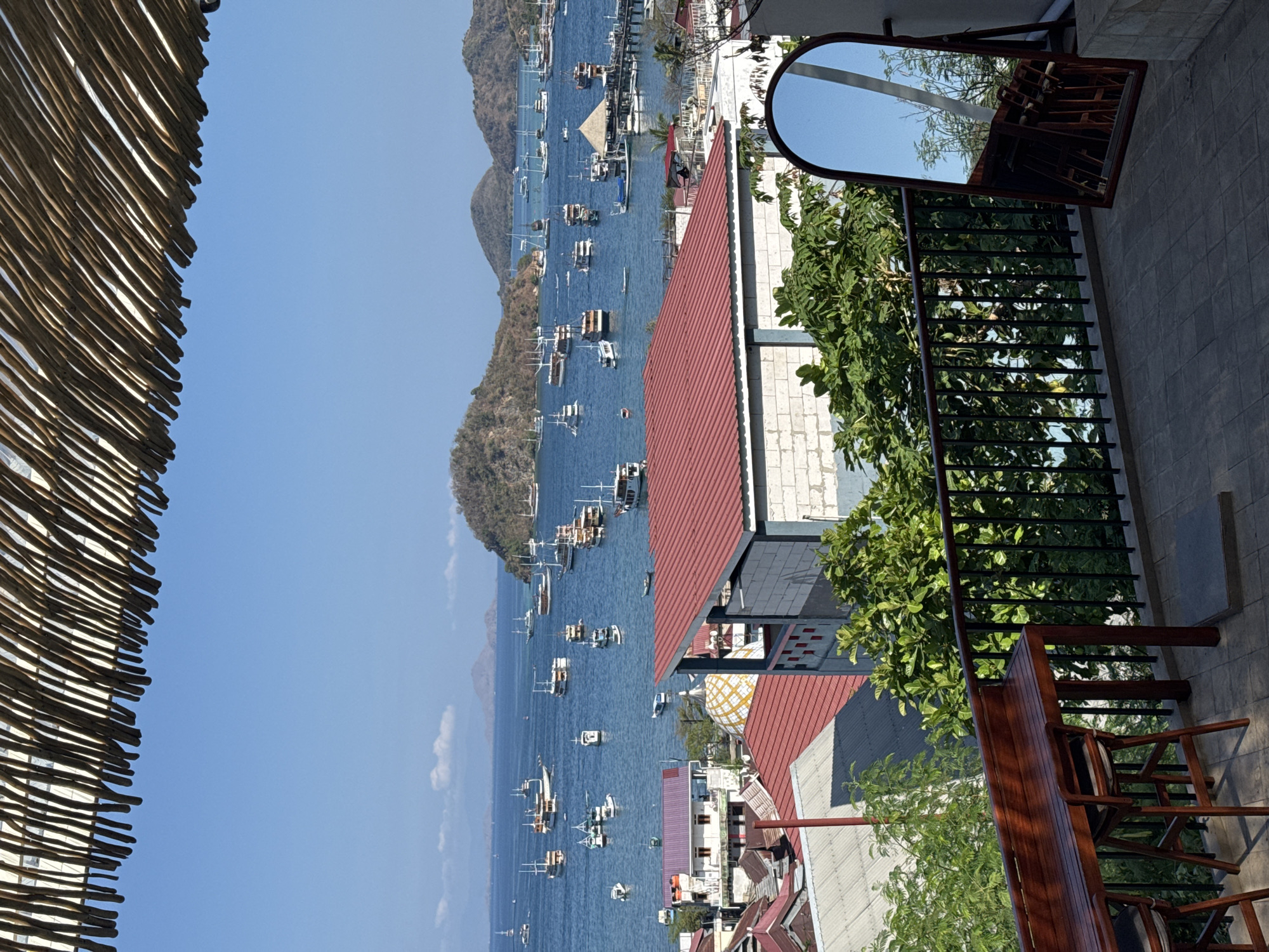 An elevated, wide-angle, exterior, daytime shot captures a vibrant harbor scene in Labuan Bajo, Indonesia, a coastal town on the island of Flores. The foreground features a rustic wooden table and chairs on a tiled balcony, with a full-length mirror positioned to the right, reflecting the scene and surrounding greenery. Beyond the balcony, a cluster of buildings with red-tiled roofs and white-washed walls are visible, indicative of the local architecture. The middle ground is dominated by the calm, blue waters of the harbor, densely populated with numerous wooden boats of varying sizes, many appearing to be traditional Phinisi schooners, moored in the bay. In the distance, lush, green, and brown hills and islands rise from the sea under a clear, bright blue sky. A bamboo-thatched awning partially frames the top of the image, adding a natural, textured element to the composition. The overall impression is one of a bustling, tropical maritime hub.