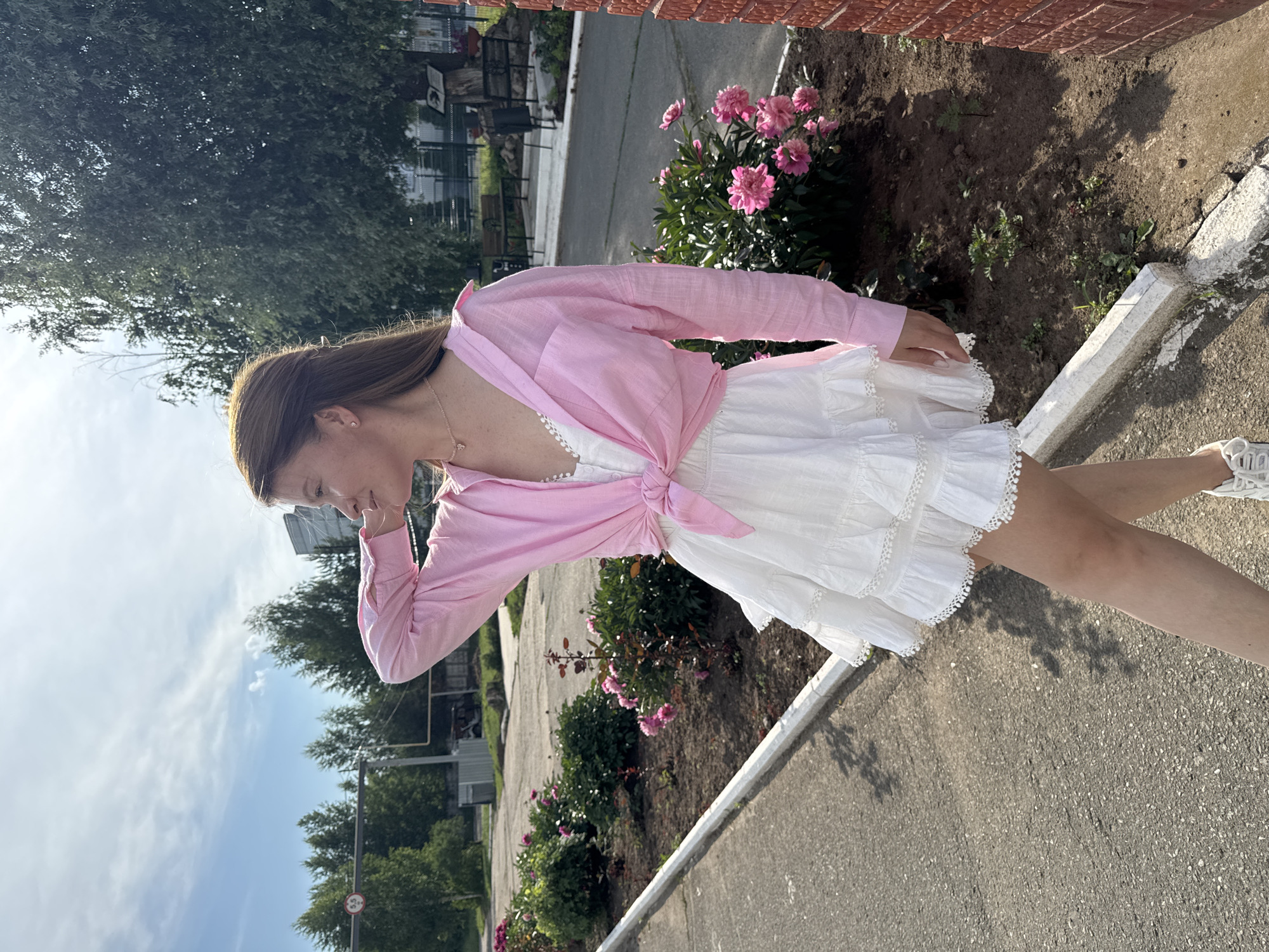 A young woman with long brown hair is standing outside in Chaikovsky, Russia, facing to her left and looking away from the camera. She is wearing a white ruffled dress and a pink unbuttoned shirt tied at the waist. Her right arm is raised, resting on her head. The background shows a sidewalk, a flower bed with pink peonies, trees, and a building. The sky is blue with scattered clouds.