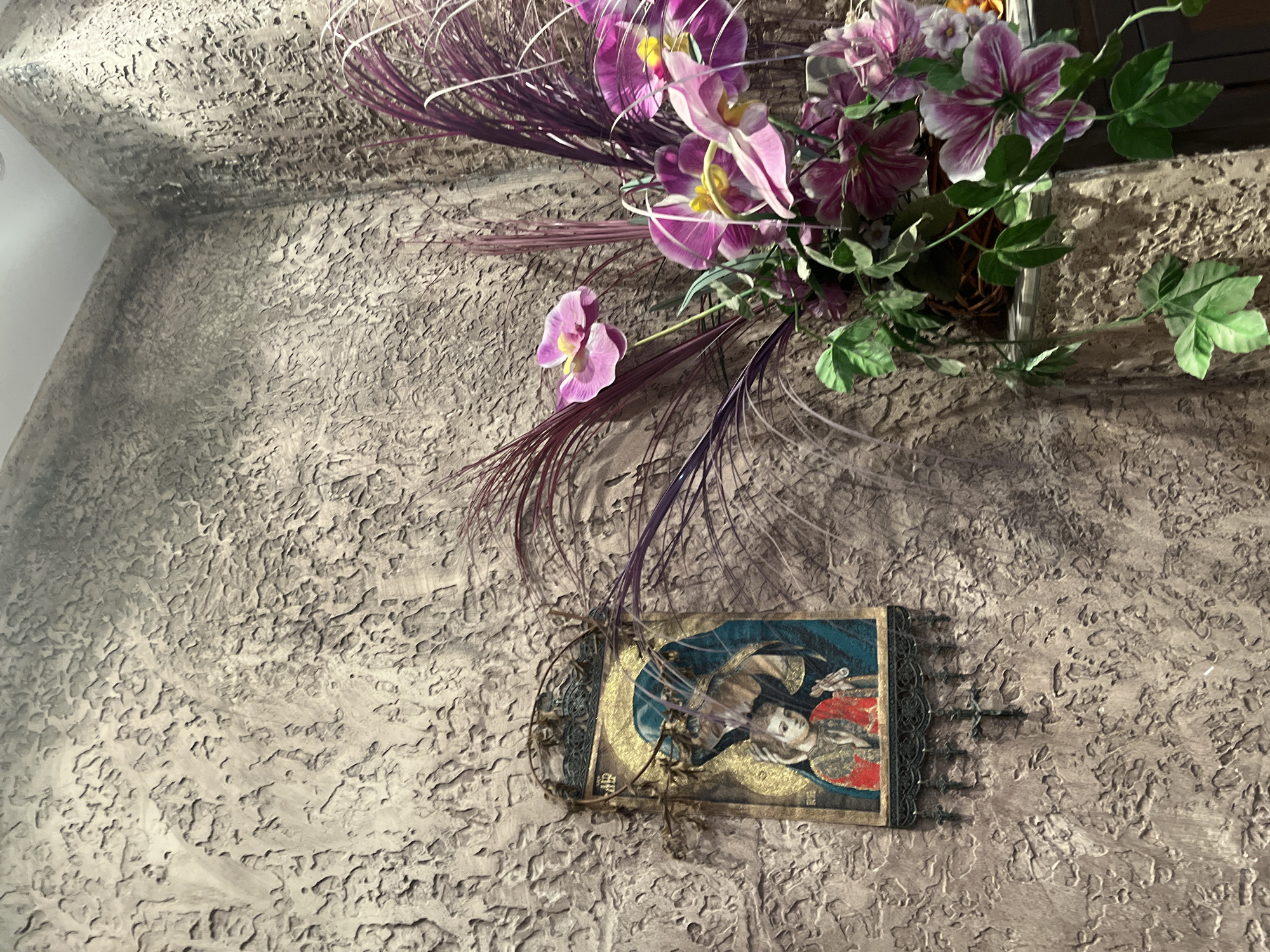 This media file is a vertical, eye-level shot of a textured, taupe-colored wall with a decorative item hanging on it. The wall has a rough, uneven surface with deep ridges and indentations, possibly stucco or a similar material. In the upper right portion of the frame, a floral arrangement is partially visible. It features artificial pink and purple orchids with yellow centers, interspersed with wispy, dark purple, feather-like foliage. The flowers appear to be in a woven basket that is mostly obscured.
Below the floral arrangement, a small, rectangular, framed item is hanging on the wall. It appears to be a religious icon or artwork. The frame has a dark, possibly metallic lace-like trim. The artwork itself depicts a figure with fair skin, light hair, and prominent eyes, facing slightly to the left. The figure is dressed in a blue garment and appears to be adorned with gold elements, possibly a halo or celestial background. A red circular object is also visible within the artwork. The overall impression is one of an indoor setting, likely a home or religious space, given the nature of the artwork. The lighting suggests an interior environment, possibly illuminated by artificial light, creating shadows on the textured wall. No specific time of day or weather conditions are discernible. The location context of Nesebar, Bulgaria, might suggest a historical or traditional setting, which aligns with the style of the icon.