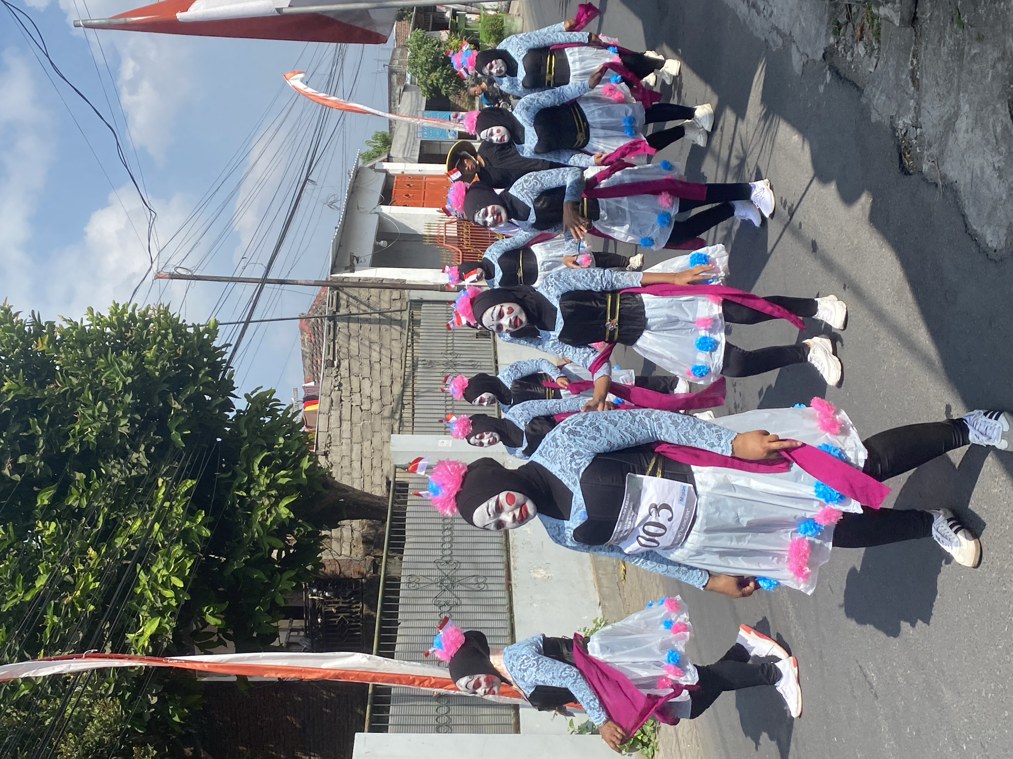 In Kediri City, Indonesia, a group of individuals, possibly participants in a parade or performance, are walking down a paved street. They are all wearing similar costumes featuring black headscarves, black and blue bodices with delicate blue lace sleeves, white skirts adorned with pink and blue pom-poms, and pink sashes. Their faces are painted with white makeup, red cheeks, and black accents, resembling clown-like or theatrical makeup. Some have pink, fluffy decorations on their headscarves. One individual in the foreground has a bib with the number "003" and text that includes "KREASI" and "M.pan." The street is lined with buildings and a low brick wall, with a large tree providing shade on the left. Indonesian flags are visible, one on a tall pole to the left and another to the right, suggesting a national celebration or event. The sky is blue with scattered clouds, and power lines are strung overhead. The overall scene depicts a sunny day with a procession moving along the street.