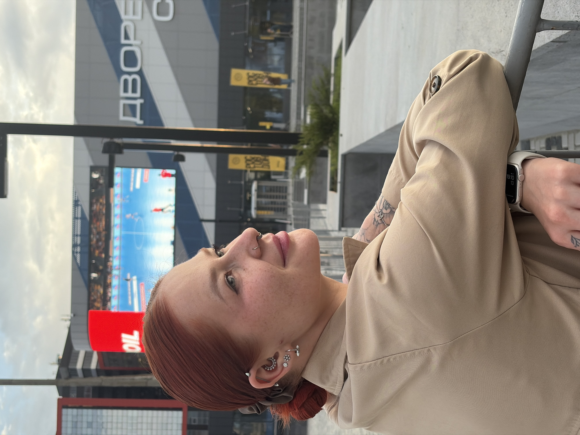 A young woman with red hair stands outside near the Rostov Arena in Rostov-on-Don, Russia. She is wearing a light brown jacket and an Apple Watch. The Rostov Arena is visible in the background, with a large screen displaying an image. The sky is overcast.
