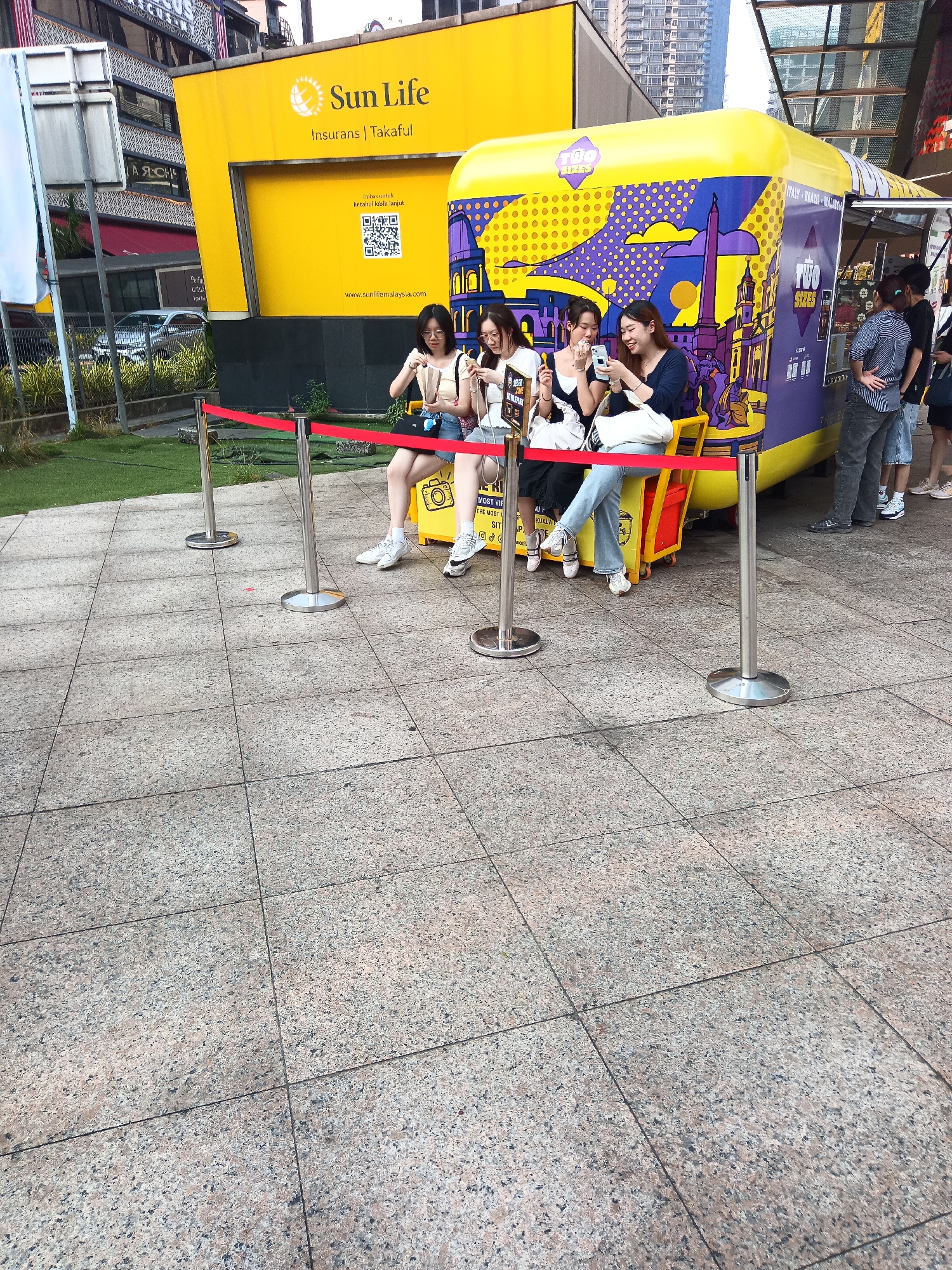 In Kuala Lumpur, Malaysia, four young Asian women are seated on a bench behind a red velvet rope barrier, each engrossed in their smartphones. They are positioned in front of a brightly colored food truck, which features purple and yellow graphics depicting European-style buildings and a sun. The food truck is adorned with the words "TWO SIZES" in a circular logo. Behind the food truck, a prominent yellow building displays the "Sun Life" logo with the tagline "Insurans | Takaful." The ground is paved with granite tiles, and a patch of green grass is visible to the left, bordered by foliage. In the background, modern skyscrapers and urban infrastructure can be seen. The overall scene suggests a lively urban setting with a promotional event or a popular food vendor.
