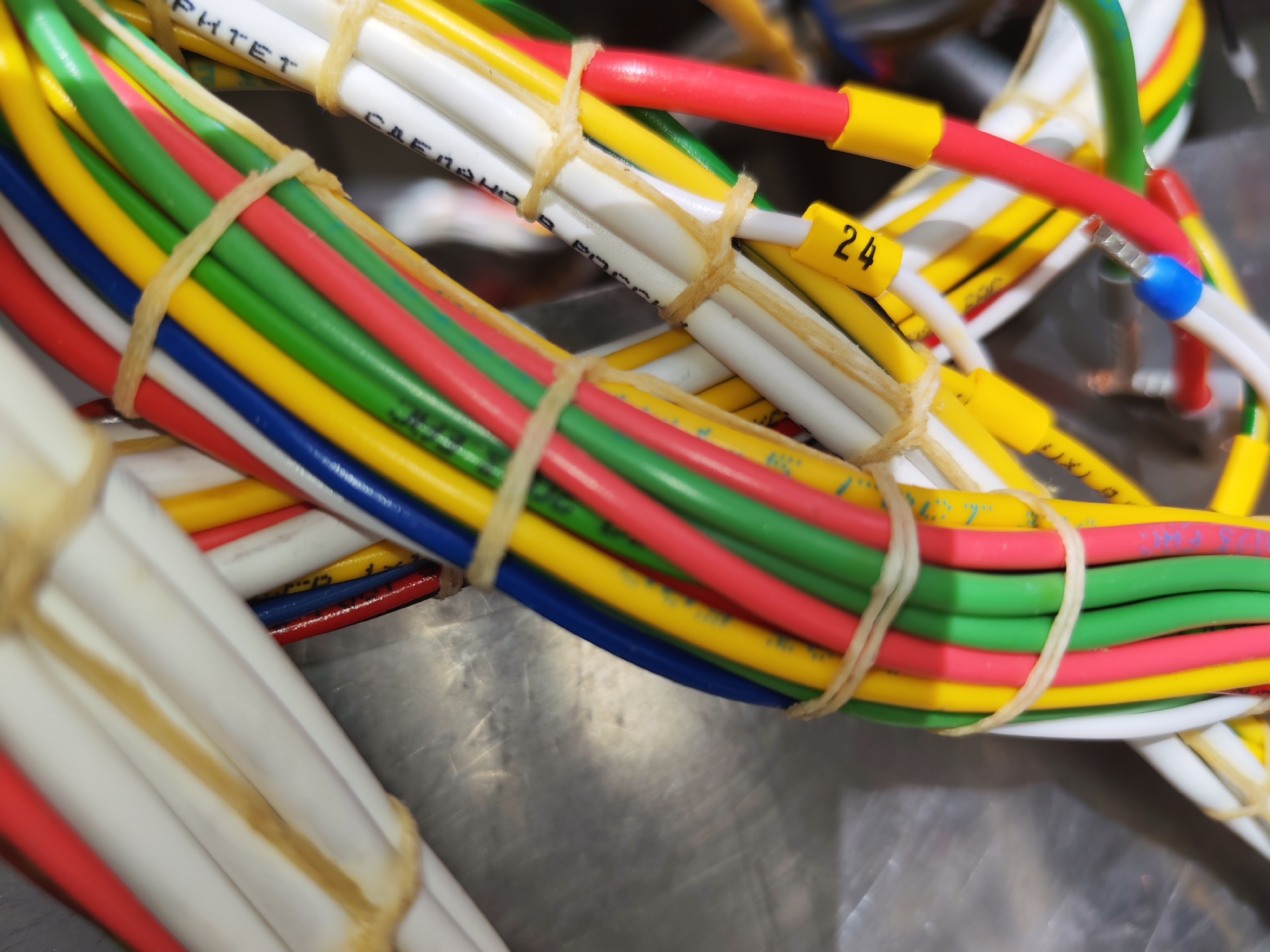 The image shows a close-up, angled view of a bundle of colored wires, primarily red, green, yellow, and blue, secured with beige elastic bands. Some of the white wires have black text printed on them, such as "CПЕЦНАЗ" and "24". There are also yellow plastic tags with numbers on some of the wires. The wires appear to be part of an electronic or mechanical system, possibly in a workshop or industrial setting. The background is metallic and slightly out of focus, suggesting a surface like a machine or a metal enclosure. There are no people or animals visible in the image. The image is a still photograph and does not depict any specific action. There is no information or context provided within the image to associate it with the city of Izhevsk, Russia, or any other specific location.