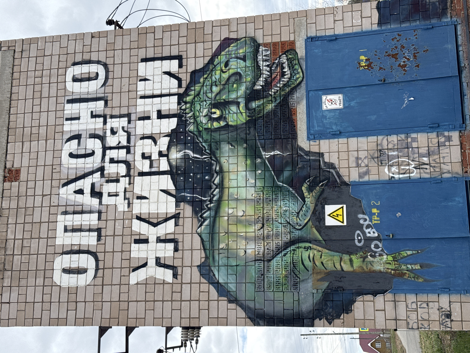 The image shows a building facade with a mural. The mural depicts a green dinosaur-like creature with its mouth open, painted on a brick wall. To the left of the creature, there is white lettering in a vertical orientation. Two blue metal doors are visible to the right of the mural, with a yellow warning sign featuring a lightning bolt on the lower door. Graffiti markings are present on both the wall and the doors. Power lines and a partially visible building can be seen in the background. This mural is located in the city of Zav'yalovo, Russia.