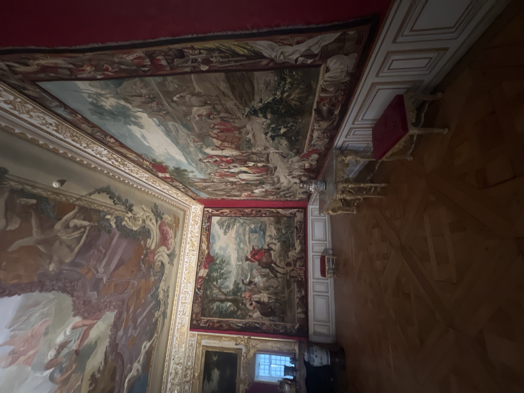This is an indoor scene in a grand, ornate room, likely part of a palace or museum, located in Oberschleißheim, Germany. The room features high ceilings with elaborate frescoes depicting mythological or allegorical scenes, rendered in soft pastel colors. The walls are predominantly a deep red, adorned with large, detailed tapestries that showcase historical or pastoral scenes with figures and horses.

The flooring is a polished parquet with a geometric pattern. The room contains several pieces of antique furniture, including two small, red-upholstered stools with gilded legs, a gilded table with ornate carvings and a white vase on it, and another table with a vase. On the left side of the image, a large, blue and white porcelain vase stands on a dark pedestal. A framed painting hangs on the wall near a window with multiple panes, letting in natural light. The architectural details include decorative moldings and panels along the lower walls, also in white and gold. The overall impression is one of opulence and historical significance.