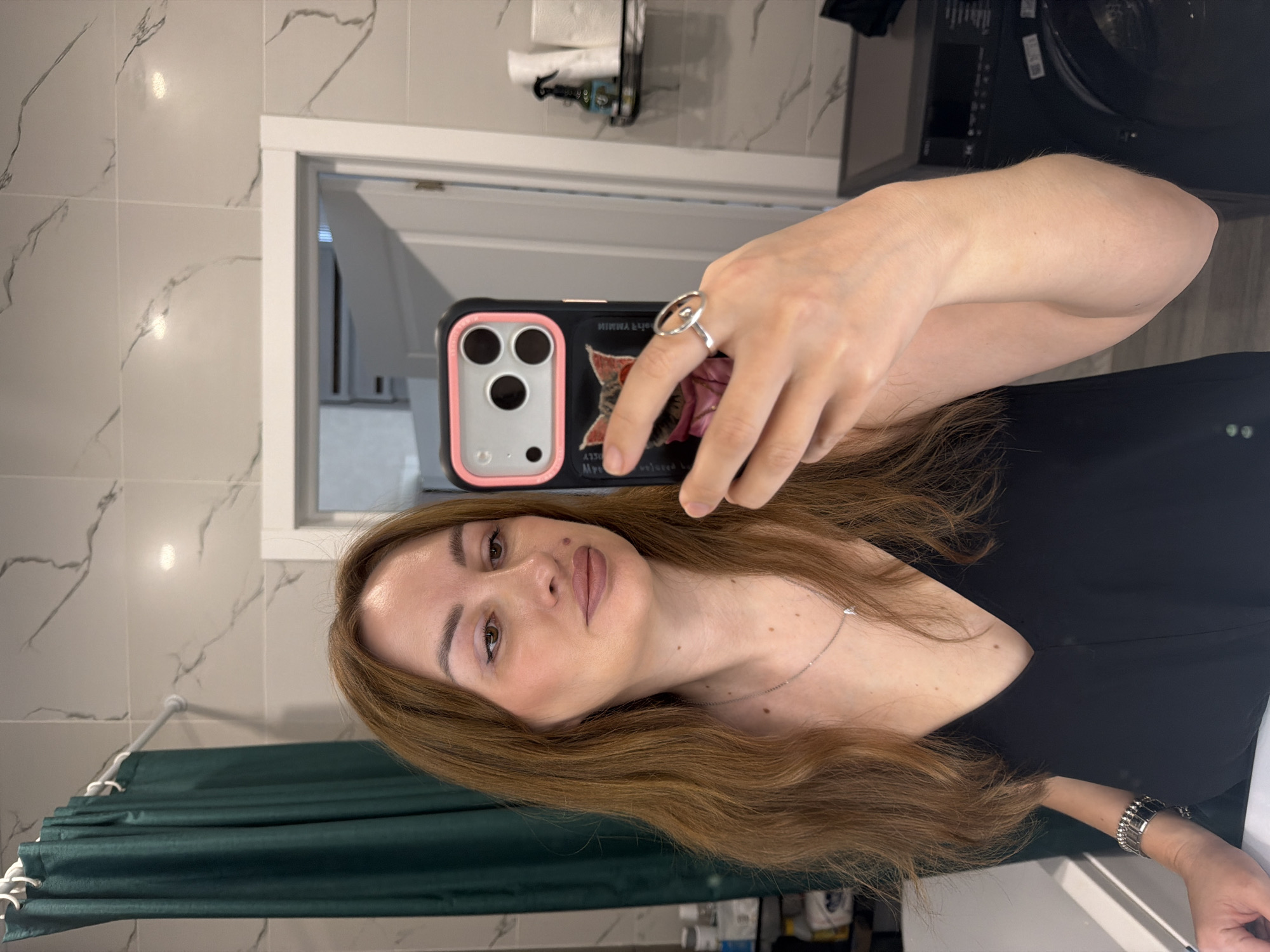 A medium shot captures a woman with long, reddish-brown hair styled loose, taking a mirror selfie in what appears to be a bathroom. She is looking at her reflection, holding a smartphone in her right hand, which has a black case adorned with a pink border and a graphic of a cat. She is wearing a low-cut black top, a thin silver necklace, and a silver ring with an oval shape on her right ring finger. A silver bracelet with black rectangular accents is visible on her left wrist. The background shows a green shower curtain on the left, white marbled tiles on the walls, and a doorway leading to another room on the right. Shelves with toiletries are visible in the background, as is a black appliance, possibly a washing machine, to the right. The lighting suggests an indoor environment.