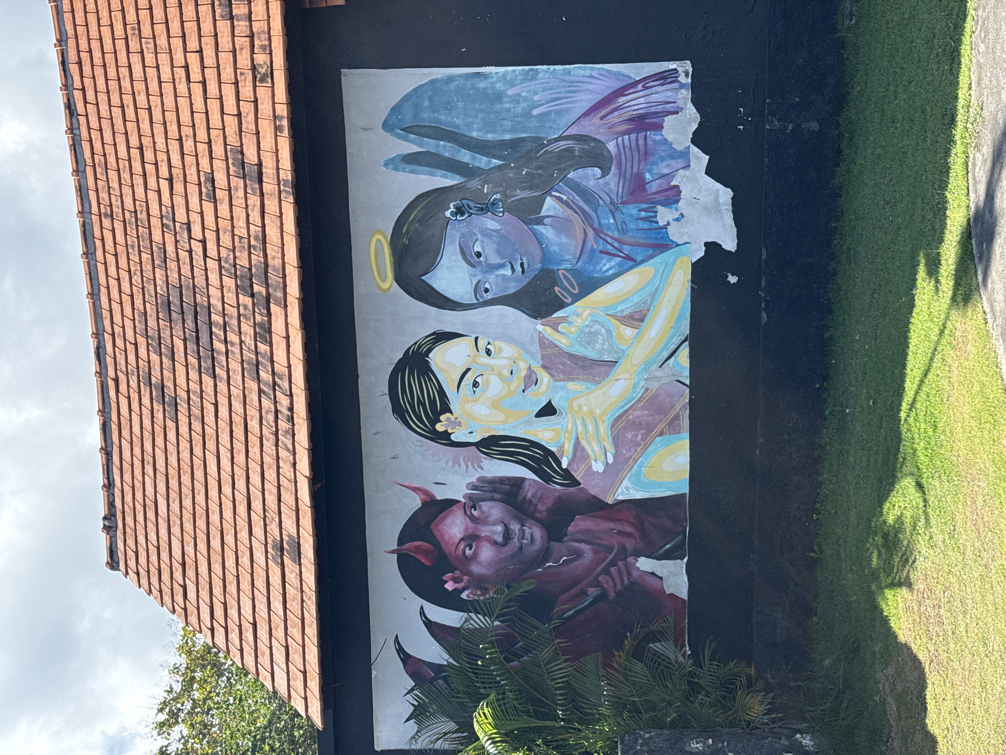 A mural is painted on the side of a building in Nusa Penida, Indonesia. The mural depicts an angel, a woman, and a devil, stacked vertically. The angel is at the top, with wings and a halo. The woman is in the middle, with her arms wrapped around the devil, who is at the bottom, complete with horns. The building has a red tile roof on the left. To the right of the mural is a patch of green grass. A sidewalk runs along the edge of the grass. Some greenery is at the bottom of the mural.