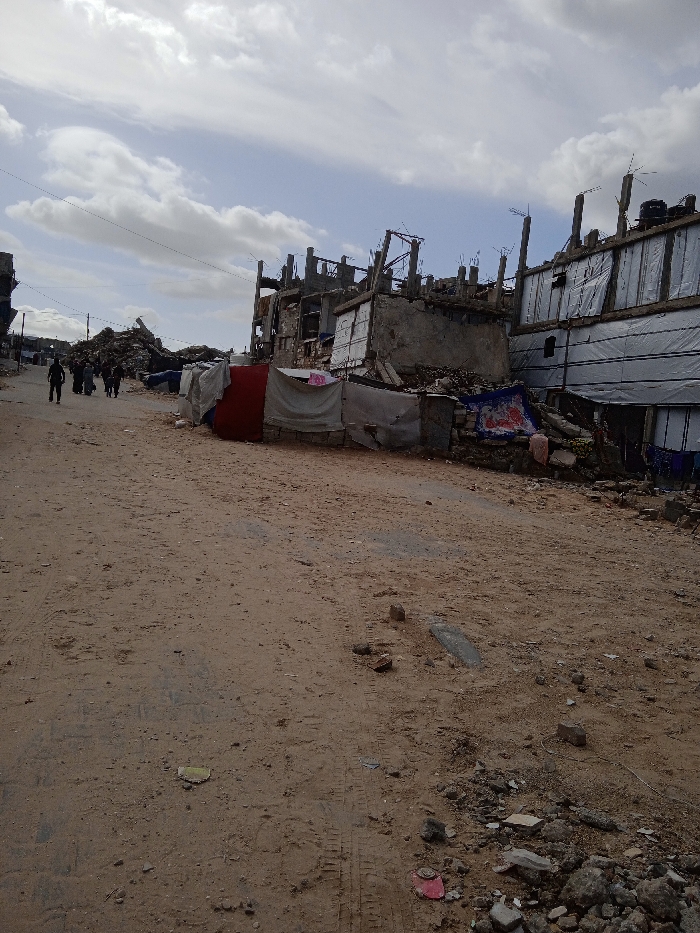 The image displays a street in Khan Yunis, Palestinian Territory, under a partially cloudy sky. The ground consists of an unpaved, light brown dirt road, marked by tire tracks and scattered with debris including rocks, concrete fragments, and small pieces of trash.
To the right, a series of severely damaged multi-story concrete buildings line the road. These structures exhibit extensive destruction, with exposed concrete frames, rebar protruding from incomplete sections, and large portions of walls missing. Several lower sections of these damaged buildings are covered with makeshift tarpaulins and fabric sheets, including white and red sections, indicative of temporary coverings or shelters. Piles of rubble and debris accumulate at the base of these structures. Antennas and what appear to be water tanks are visible on the rooftops of some damaged buildings.
In the mid-distance on the left, a substantial mound of building debris and collapsed structures marks the extent of further destruction. A group of approximately five to seven individuals, dressed in dark clothing, are walking away from the camera on the dirt road, heading towards the distant rubble. Utility lines are visible suspended across the upper left portion of the frame.
