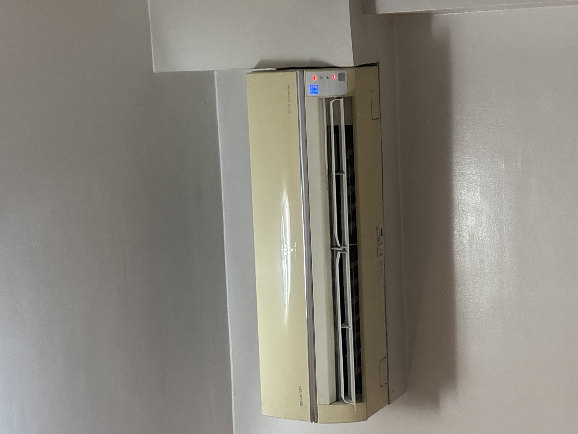 The image shows a close-up, eye-level view of a light beige Sharp air conditioning unit mounted on a light gray wall, likely in an indoor setting. The air conditioning unit is the primary subject, filling the majority of the frame. It has a horizontal vent at the bottom with adjustable louvers and a digital display panel on the right side showing a blue screen with what appears to be temperature settings and several indicator lights, some of which are illuminated red. The text "SHARP" is visible on the left side of the unit, and "ECO Inverter" is printed on the right. The wall has a slightly textured finish and is painted a neutral, light color. The ceiling is visible at the top of the image, also a light color. The lighting in the scene appears to be artificial, casting a soft glow on the surfaces. There are no people or animals present in the image. The overall scene suggests a typical residential or commercial interior space. Given the context of Pasay City, Philippines, this could be a room in a hotel, condominium, or office building.
