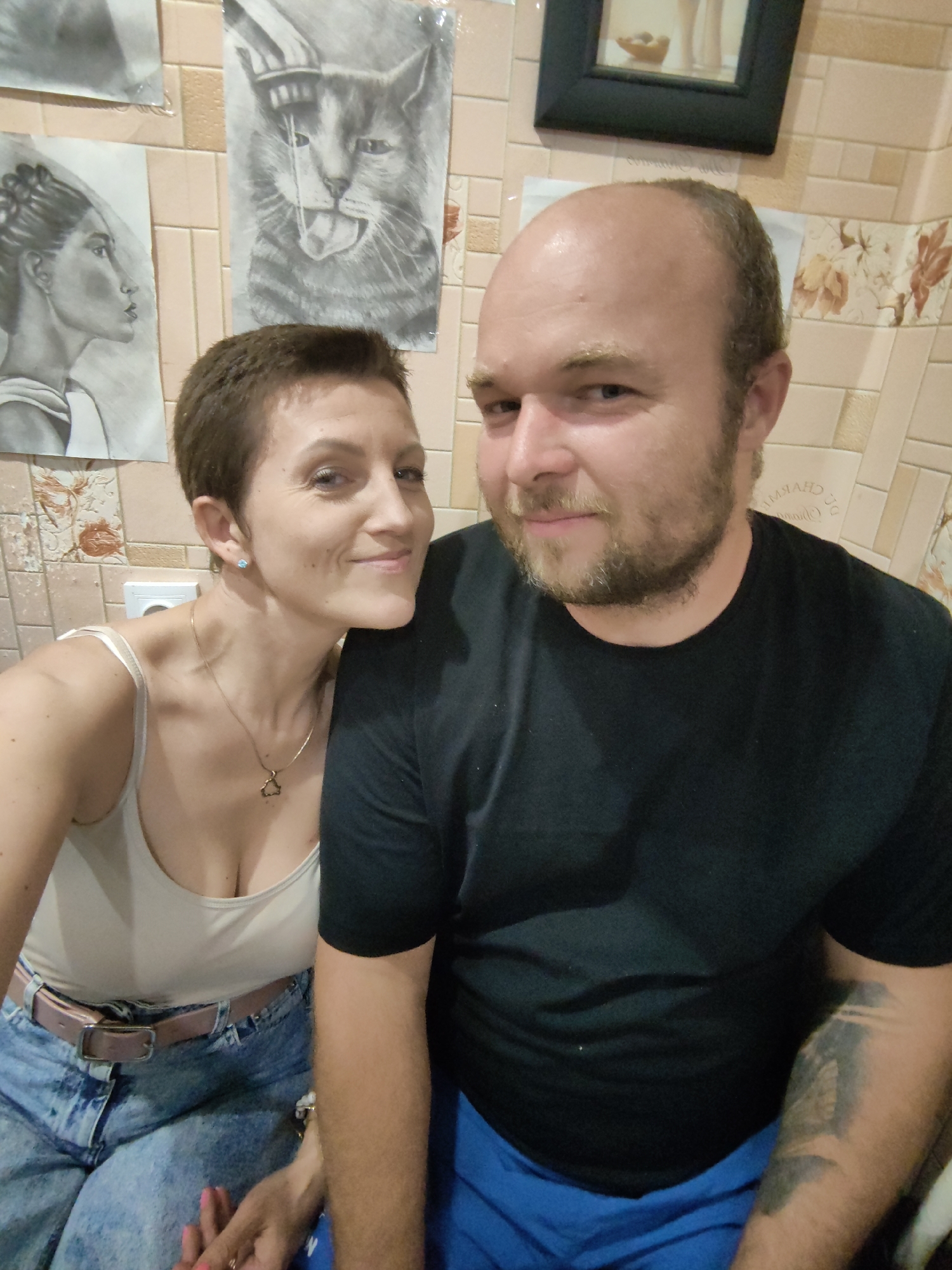 A medium shot photograph captures a man and a woman in a close-up pose, with the woman leaning her head on the man's shoulder and looking towards the camera with a smile. They are positioned in what appears to be a domestic setting, possibly an apartment in Myadzyel, Belarus, as indicated by the tile pattern on the wall that matches known decorative styles in the region.
The woman, on the left, has short, dark brown hair and is wearing a light-colored tank top with spaghetti straps and a gold necklace. A pink and white belt cinches her high-waisted denim jeans. Her fingernails are painted pink, and she is wearing a ring on her left hand. She has a light blue earring in her visible ear.
The man, on the right, is facing the camera with a slight smile. He has a bald head with some light brown stubble on his chin and cheeks. He is wearing a black t-shirt and blue shorts. A tattoo is visible on his right forearm.
In the background, several framed drawings are mounted on the tiled wall. One drawing depicts a cat with its tongue hanging out, another is a profile of a woman's face, and a third, higher up, is a framed picture of a vase with some fruit. A white electrical outlet is visible behind the woman's head. The overall lighting is soft, suggesting indoor illumination.