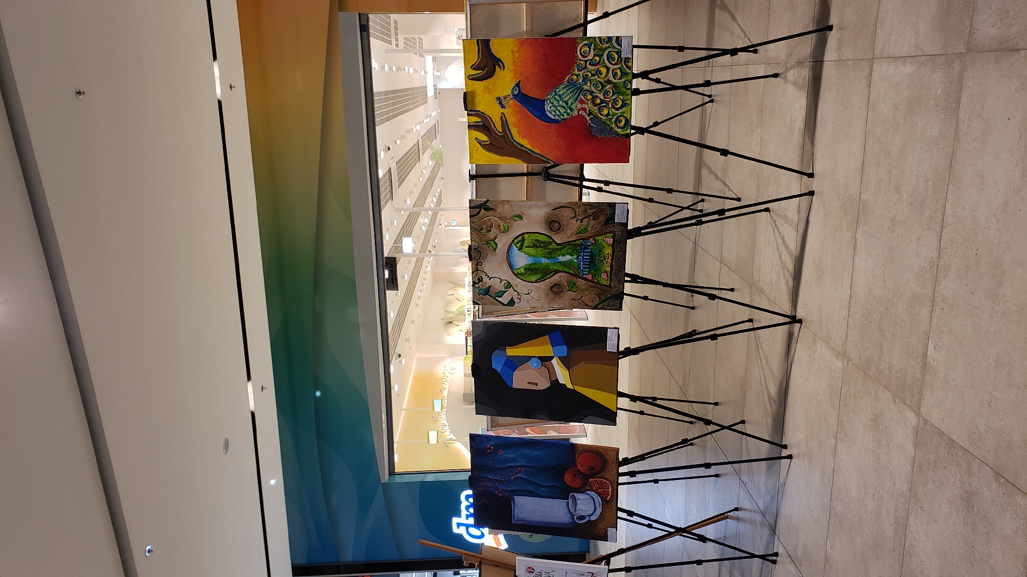 Four paintings are displayed on easels in a modern indoor public space. From left to right, the first painting depicts a still life with a white vase, two pomegranates, and a sliced orange. The second painting is a stylized portrait of a figure with a blue headscarf and yellow garment, reminiscent of "Girl with a Pearl Earring." The third painting shows a landscape viewed through a keyhole shape, with greenery and a waterfall. The fourth painting features a peacock with iridescent feathers against a vibrant sunset-colored background, with tree branches in the foreground. Behind the paintings, a large storefront with the "dm" logo is visible, indicating a retail environment. The floor is tiled, and the ceiling is modern with recessed lighting. The overall setting appears to be a shopping mall or a similar public exhibition space. The paintings are likely part of an art exhibition or display. The location is identified as the OC MAX shopping center in Prešov, Slovakia. There are no people visible in the frame.