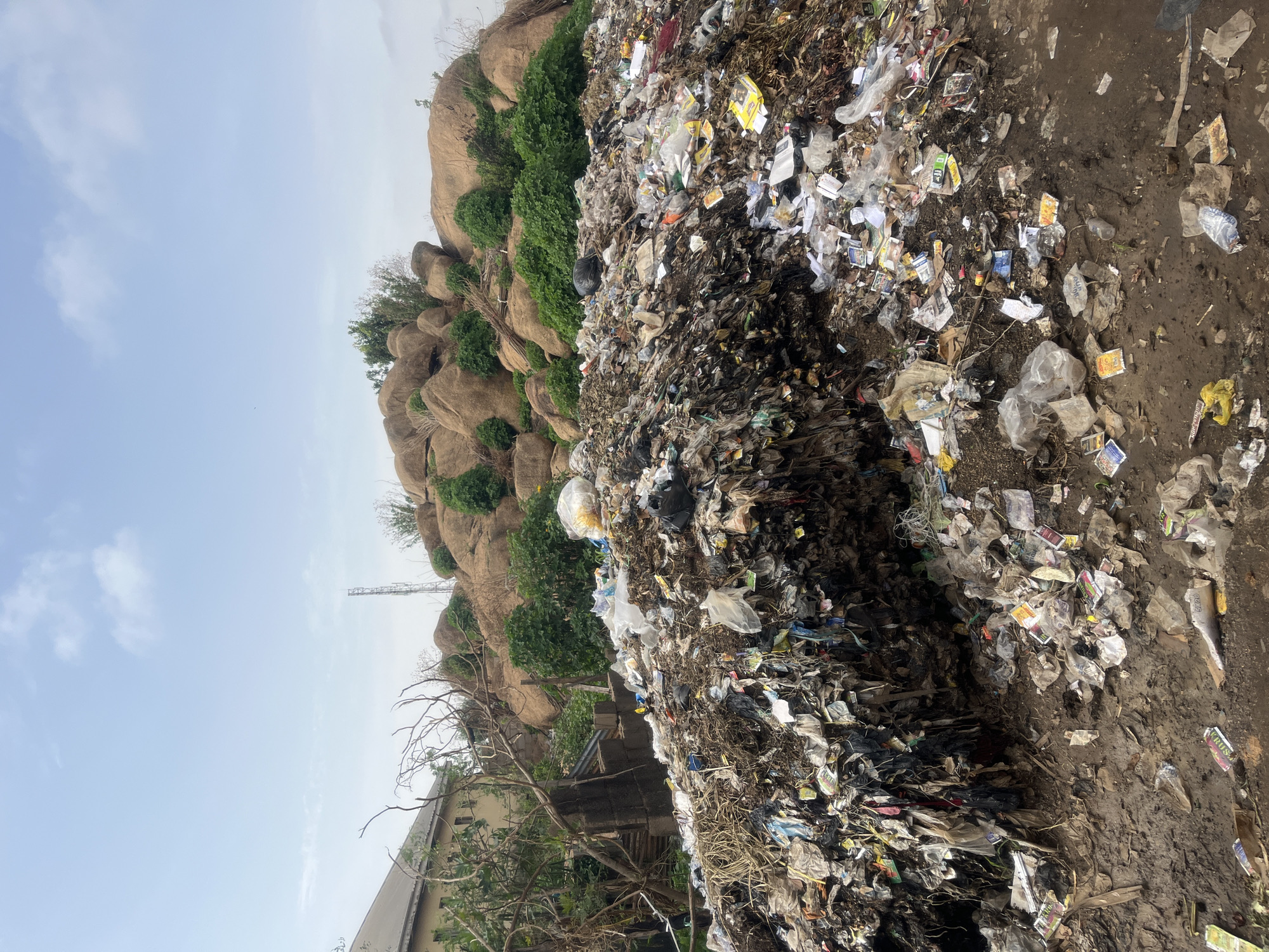 A large pile of garbage is in Gura Top, Nigeria. The garbage consists of plastic bags, paper, and other debris. A rock formation covered in green bushes rises in the background. The ground is dirt and mud, and the sky is overcast. A building and a communication tower can be seen behind the garbage pile.