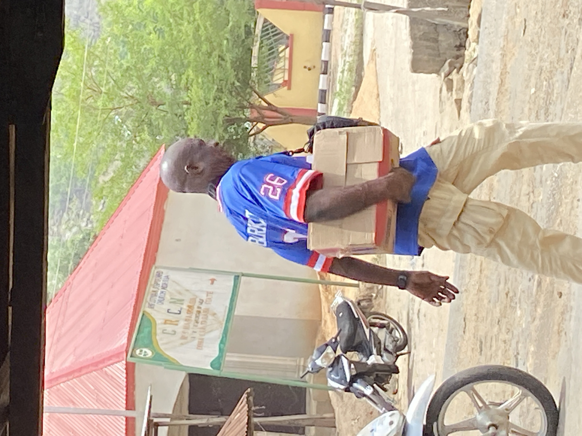 In Mararaba, Nigeria, an adult male carries several cardboard boxes. He is wearing a blue shirt with white and red trim, and the number "26" is visible on the back. He is also wearing light-colored pants. Behind him, there is a building with a red roof, and a sign that reads "CRCN", partially obscuring further text. A motorcycle is parked nearby. A tree is in the background.