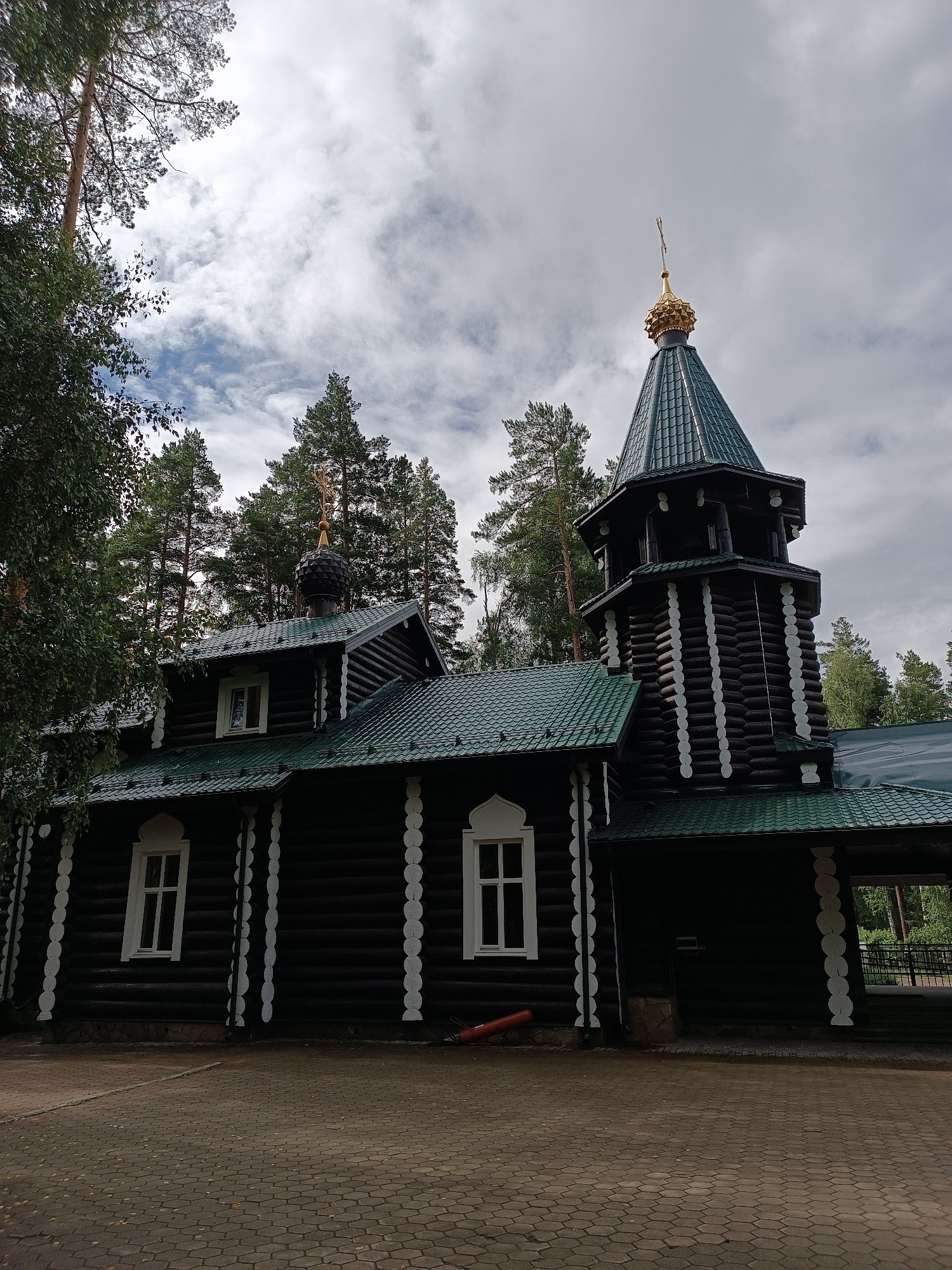 A dark wood log church with a green metal roof and a gold-domed steeple stands under a cloudy sky, surrounded by tall pine trees. The church has white trim around its windows and decorative white elements along its exterior walls. The ground in front of the church is paved with interlocking hexagonal stones. The scene appears to be a serene, natural setting, likely outside of a bustling urban area. This building is located in the city of Yekaterinburg, Russia.