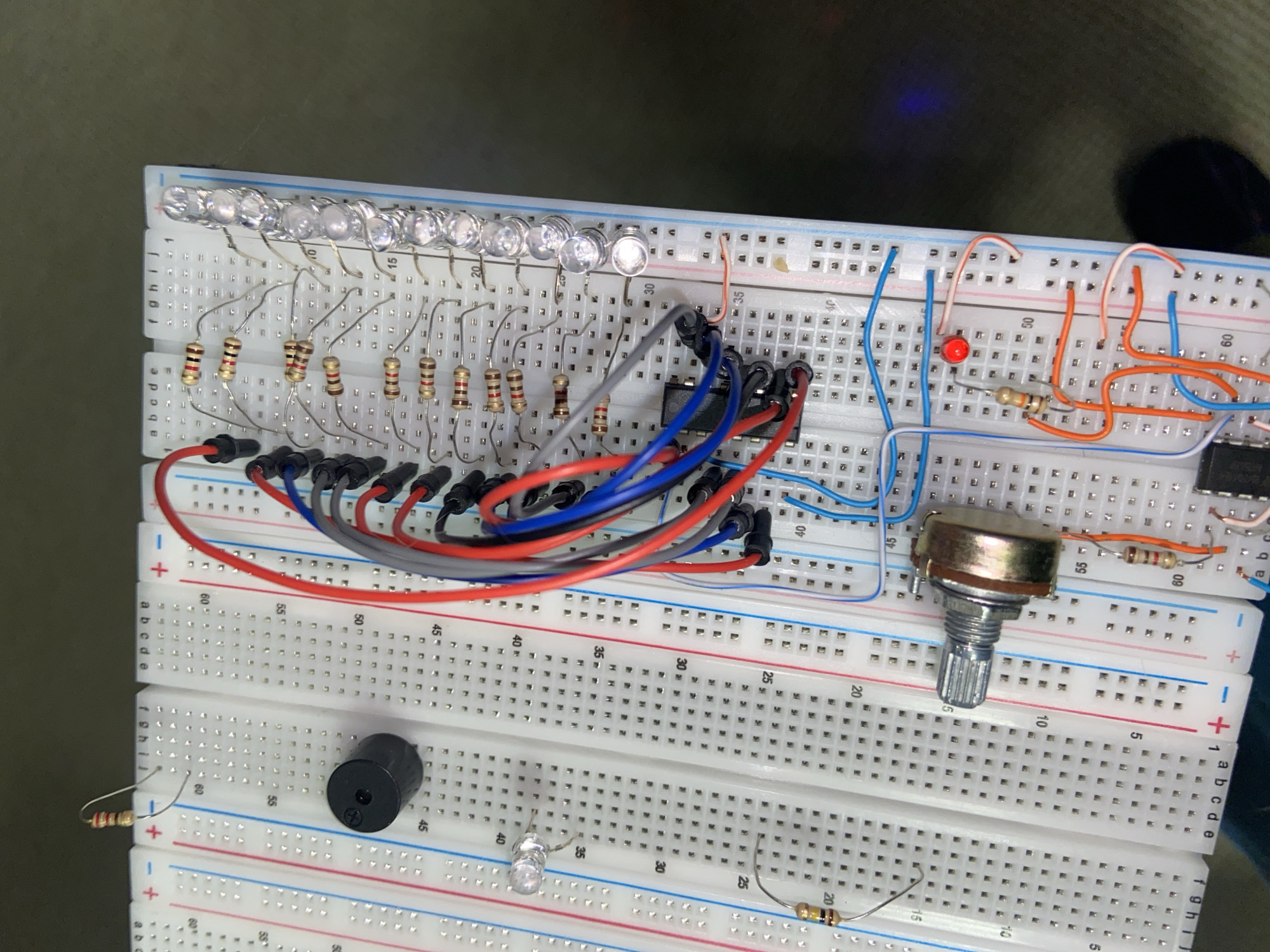 In an unknown, dimly lit indoor environment, a breadboard setup for an electronic circuit is the primary subject. The breadboard, a white plastic board with numerous holes, is covered with various electronic components and jumper wires.
Prominently visible are several clear LED lights arranged in a line along the right edge of the breadboard, connected to a series of resistors. A black buzzer component is situated on the left side, also connected via wires. Other components include a potentiometer (a knob with a metal shaft) and multiple resistors of varying colors, likely indicating different resistance values.
The circuit is connected using a multitude of colored jumper wires, including red, blue, grey, and orange, which bridge connections between the components on the breadboard. A small black integrated circuit (IC) is also present, with several wires connected to its pins. A single red LED is lit, suggesting some part of the circuit is active.
The background is dark and out of focus, with hints of a dark surface and a small blue light, possibly from another electronic device. The perspective is a top-down view, focusing on the complexity of the electronic components and their arrangement on the breadboard. There are no people or animals visible in the image. The scene appears to be a workspace for an electronics project.