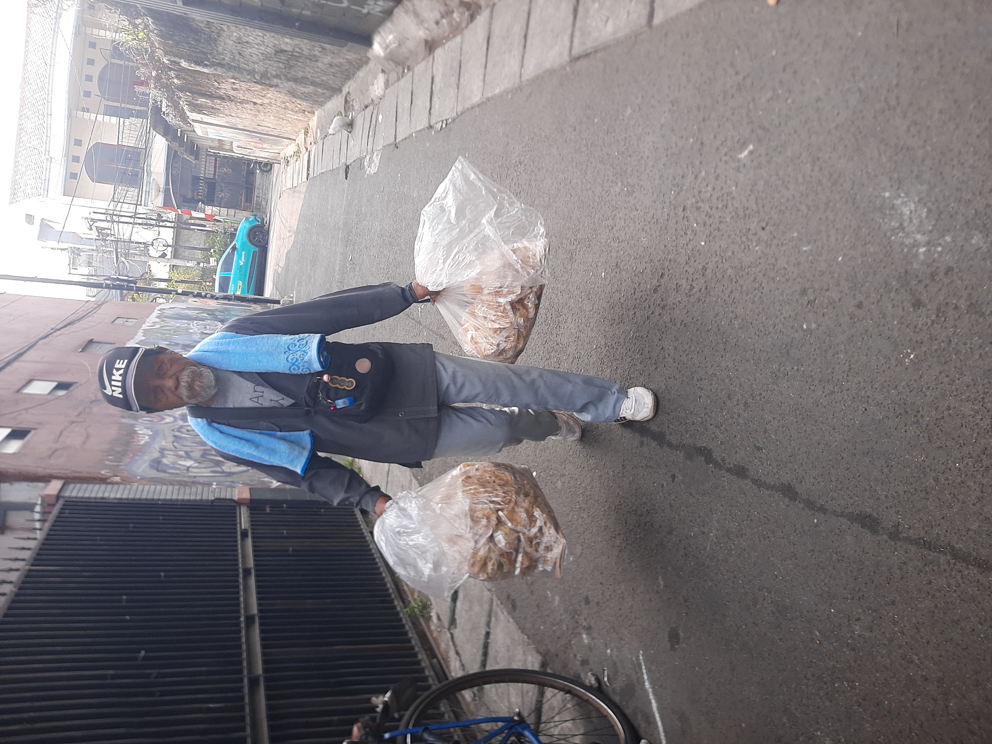 An elderly man with a grey beard, wearing a black Nike cap, a grey long-sleeved shirt, grey pants, and white sneakers, walks on an asphalt road in Central Jakarta, Indonesia. He carries two large clear plastic bags filled with food items. A blue towel is draped around his neck. He has a small black bag with items hanging from his shoulder. Beside him on the left is the rear wheel of a blue bicycle. The road is bordered by a tall black metal fence on the left and a concrete wall on the right. Buildings and power lines are visible in the background. A small white cat is visible on the right side of the road. A turquoise car is parked further down the road.