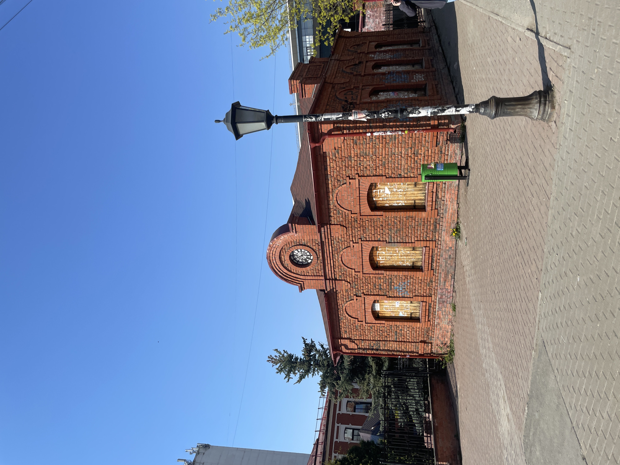 The image depicts a two-story brick building in Izhevsk, Russia, located next to a sidewalk. The building's windows are boarded up, and there is a round clock face above the entrance. A street lamp stands in front of the building, and a green trash can is positioned nearby. The sky is clear and blue. Other buildings and greenery are visible in the background.