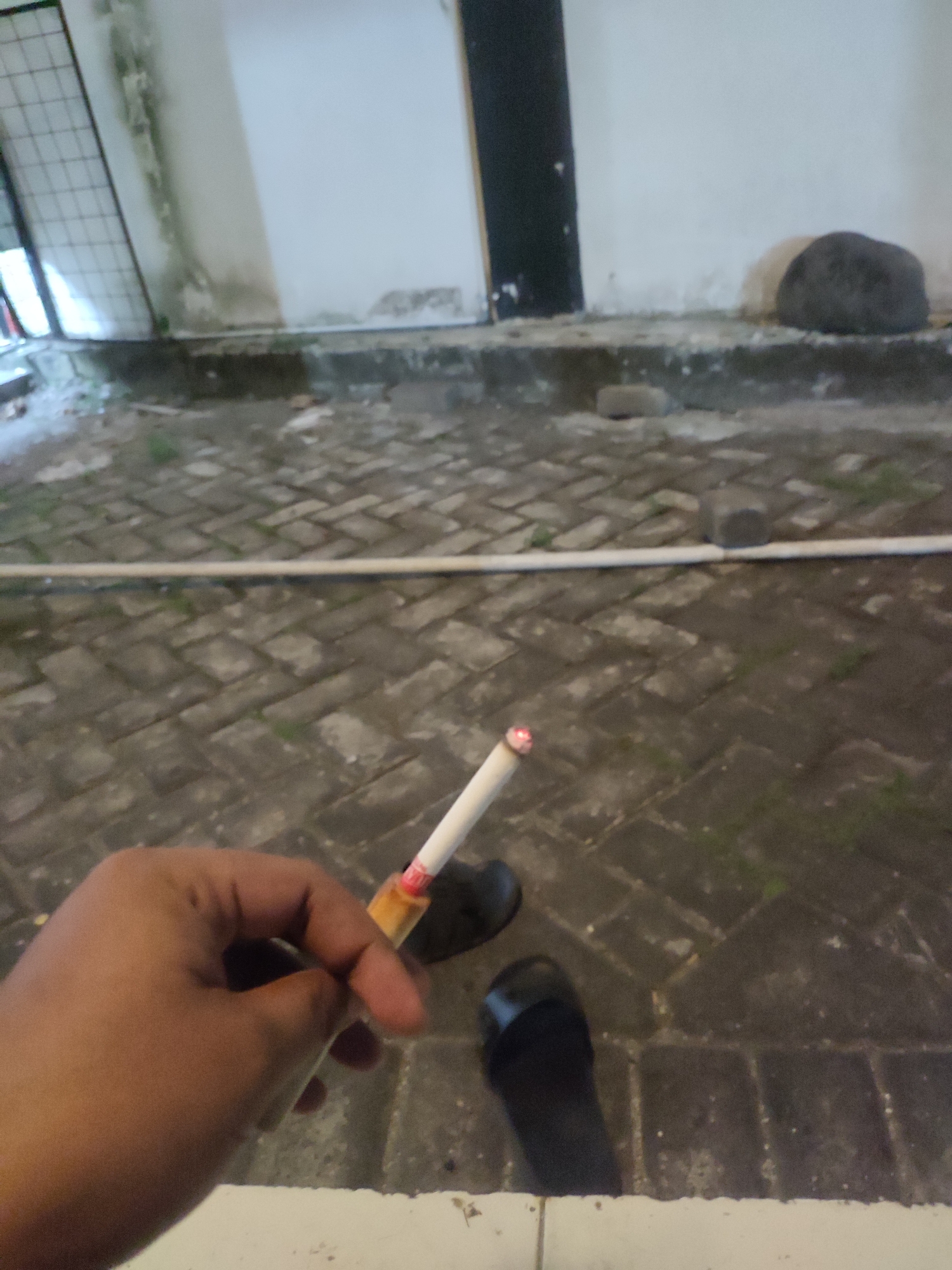 In Jember, Indonesia, a person's hand is holding a lit cigarette. The cigarette has a white filter and a brown tobacco end with a glowing red ember. The hand is a medium-toned skin color and appears to be grasping the cigarette between the thumb and index finger. The background consists of a paved area with interlocking dark gray bricks, and a white pipe runs horizontally across the middle ground. To the left of the pipe, a metal grate is visible, and beyond that, a building with white walls is present. There are also two dark-colored flip-flops or sandals visible on the paved ground. In the distance, two dark, rounded objects that appear to be stones or small boulders are positioned near the base of the white wall. The overall lighting suggests it might be dusk or night.