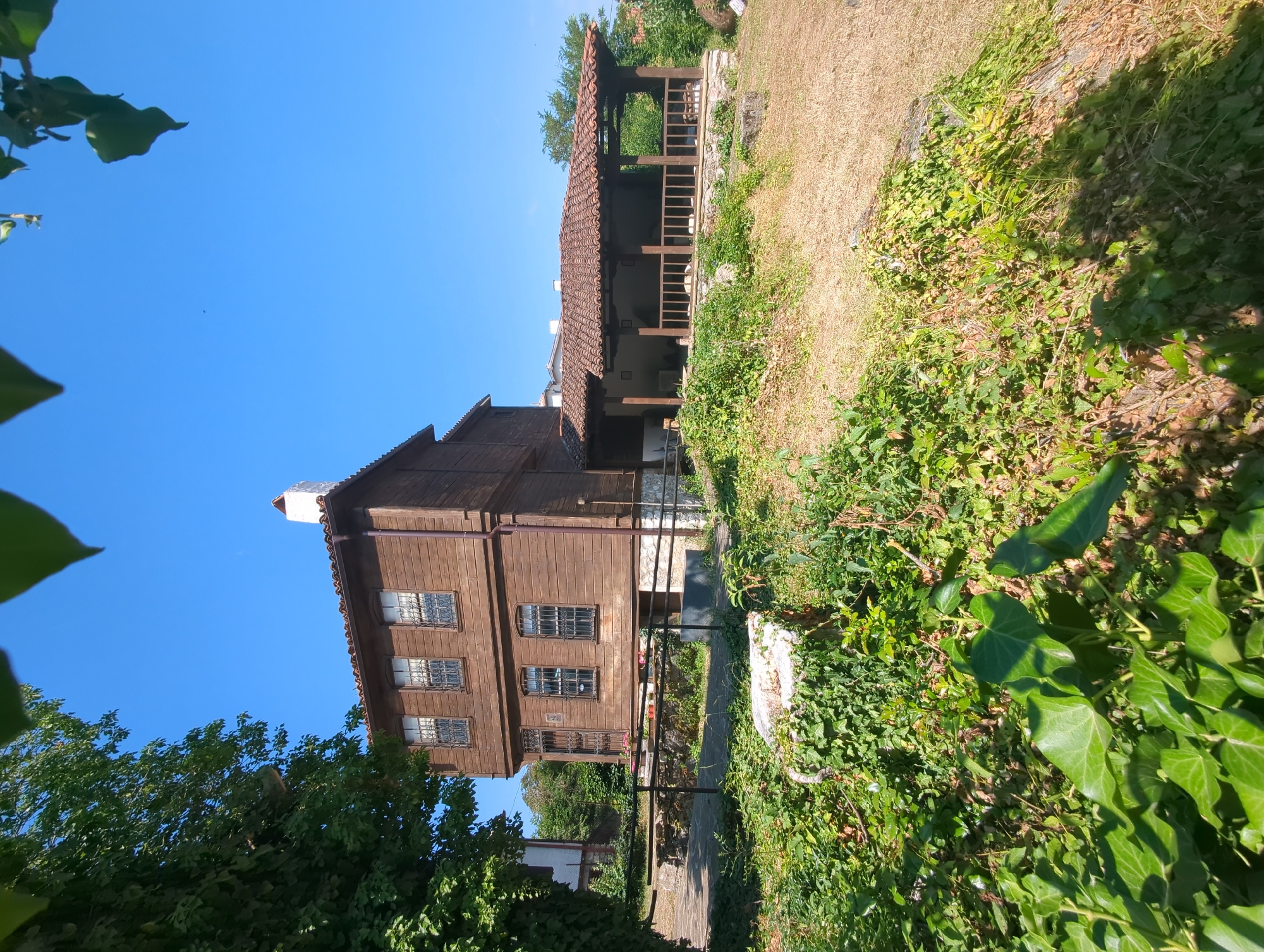 The image shows a wooden house with a tiled roof and a porch attached. The house has several windows with bars. The building is surrounded by greenery, with trees and ivy growing around it. The sky is clear and blue. This is a view of the Stoil family house in Malko Tarnovo, Bulgaria, which is a museum.
