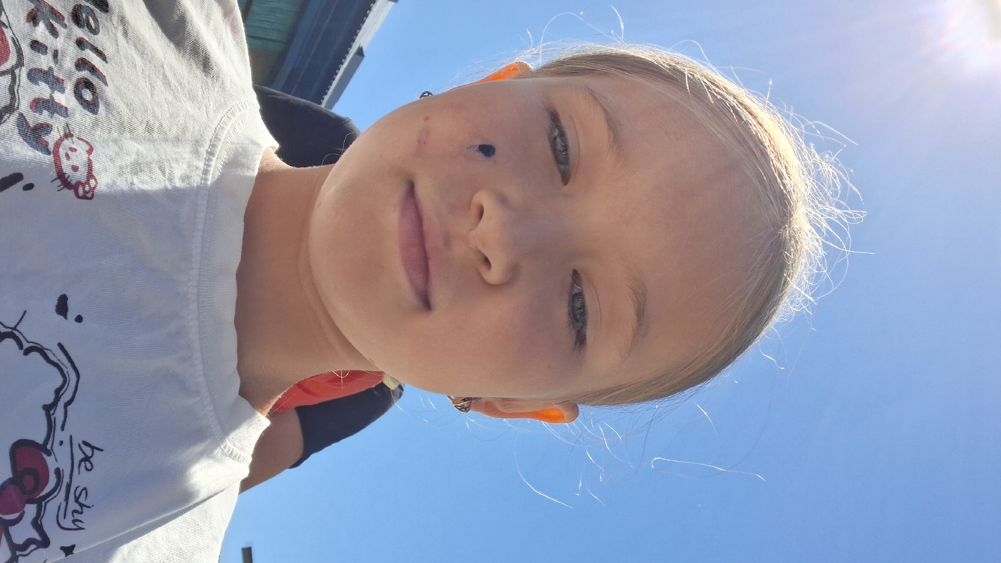 A young girl with blonde hair tied back, wearing a white t-shirt with a "Hello Kitty" design, is captured in a close-up, low-angle selfie. She has a black mark on her left cheek and a small mark or scratch below it. She is looking down and to her right. The background is a clear blue sky with a bright sun flare in the upper left corner. Behind her, a building with a dark roofline and possibly wooden siding is visible, along with blurred figures of other people. The image does not contain enough visual information to determine the specific location within the city of Semey, Kazakhstan.