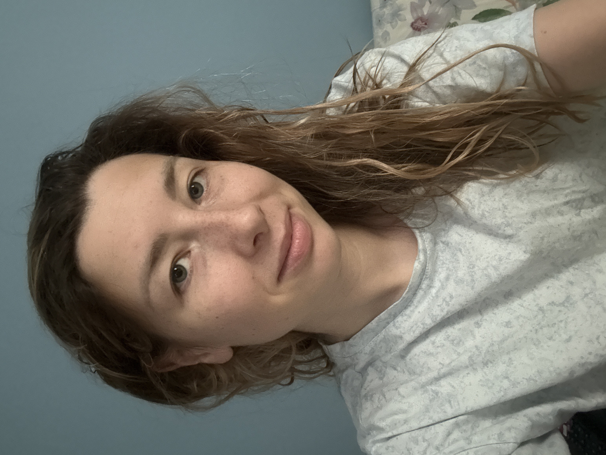 A close-up, medium shot shows a woman with light skin, freckles, and light brown hair against a blue wall. She is wearing a light gray t-shirt with a speckled pattern. Her hair is wavy and falls over her shoulders. She is looking at the camera with a slight smile. There is a floral pattern visible on the right side of the image, suggesting the presence of furniture or bedding. The lighting is soft and natural. The image does not contain any information that would allow for the determination of its location in Moscow, Russia.