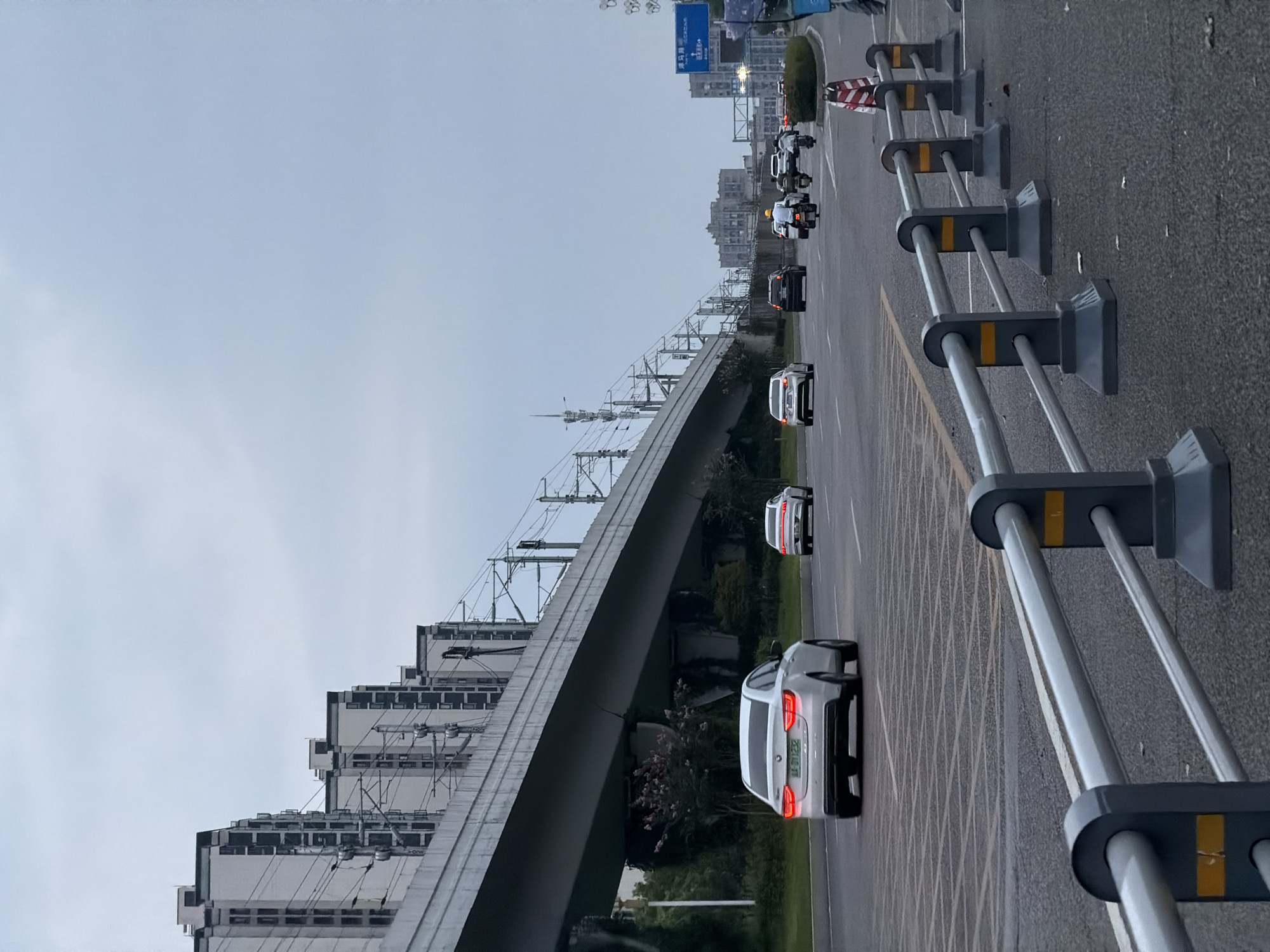 This is a medium-shot, eye-level, vertical photograph of a multi-lane road in Jiangjin, China, under an overcast sky. A section of elevated infrastructure, likely a light rail or monorail track, stretches across the upper portion of the image, supported by concrete pillars. The track is lined with overhead power lines and supporting poles.
On the road, several vehicles are in motion. In the foreground lane, a white sedan with its taillights illuminated is traveling away from the camera. Behind it, another white sedan is also visible. Further down the road, a black car and several motorcycles are present. A person on a motorcycle wearing a yellow helmet is prominent among them.
The roadside is bordered by a grey metal guardrail with yellow markings. Beyond the road, there are green trees and bushes. In the background, a row of modern, multi-story residential buildings with white facades and dark-trimmed windows are visible, receding into the distance. A blue road sign with white text and symbols is also present on the right side of the image. The overall scene depicts urban transportation infrastructure and daily life on a cloudy day.