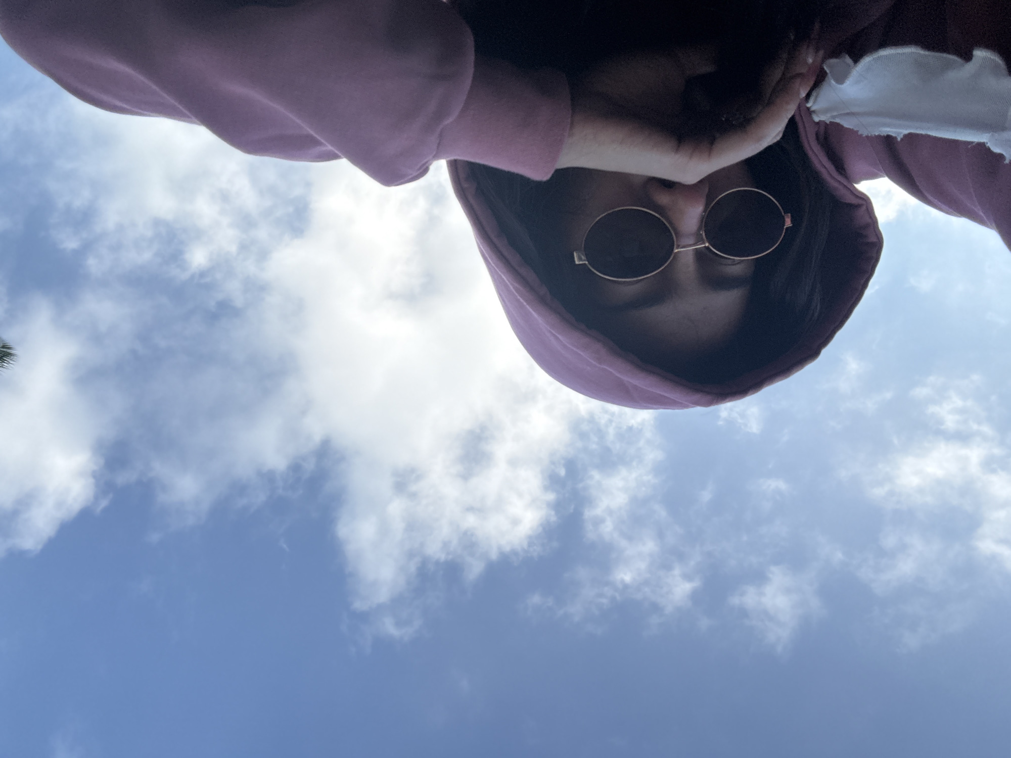 A low-angle, upward-looking shot captures a person against a bright blue sky dotted with scattered white clouds. The subject wears a pink or purplish hooded sweatshirt, with the hood pulled up. Round, dark-lensed sunglasses with thin gold frames obscure their eyes. A hand is positioned near their right temple, seemingly holding the hood or adjusting their hair. A flash of white fabric is visible near their right shoulder, peeking from beneath the sweatshirt. In the lower left corner, a dark green shape, possibly a palm frond, indicates an outdoor setting. The image is taken during the daytime under clear, sunny conditions with some cloud cover. No text is visible.