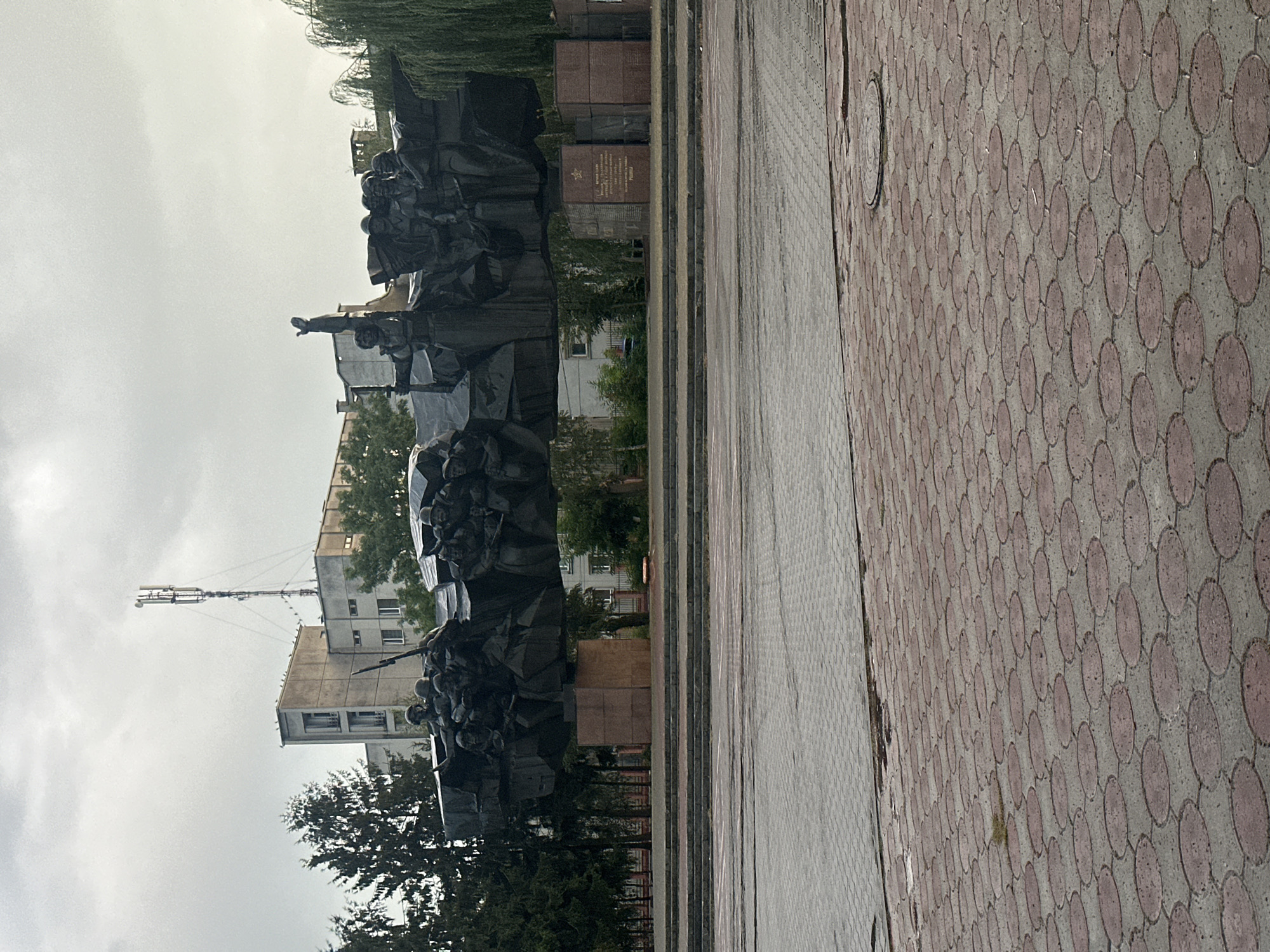 The image depicts a war memorial in Khmilnyk, Ukraine. A large, dark-colored, abstract sculpture composed of numerous figures, likely soldiers, is the central focus. The sculpture appears to be made of metal and is elevated on substantial stone pedestals. Behind the sculpture, several multi-story buildings with neutral-colored facades are visible, some with visible windows and balconies. A tall telecommunications tower with antennae rises above one of the buildings. Evergreen trees flank the sculpture on the left, and weeping willow trees are on the right. The ground in front of the memorial features tiered steps and a paved area with intricate patterns of red and gray interlocking circular and square tiles. The sky is overcast, suggesting a cloudy or possibly rainy day. The overall scene is static, with no people or discernible activity shown in the immediate vicinity of the memorial.