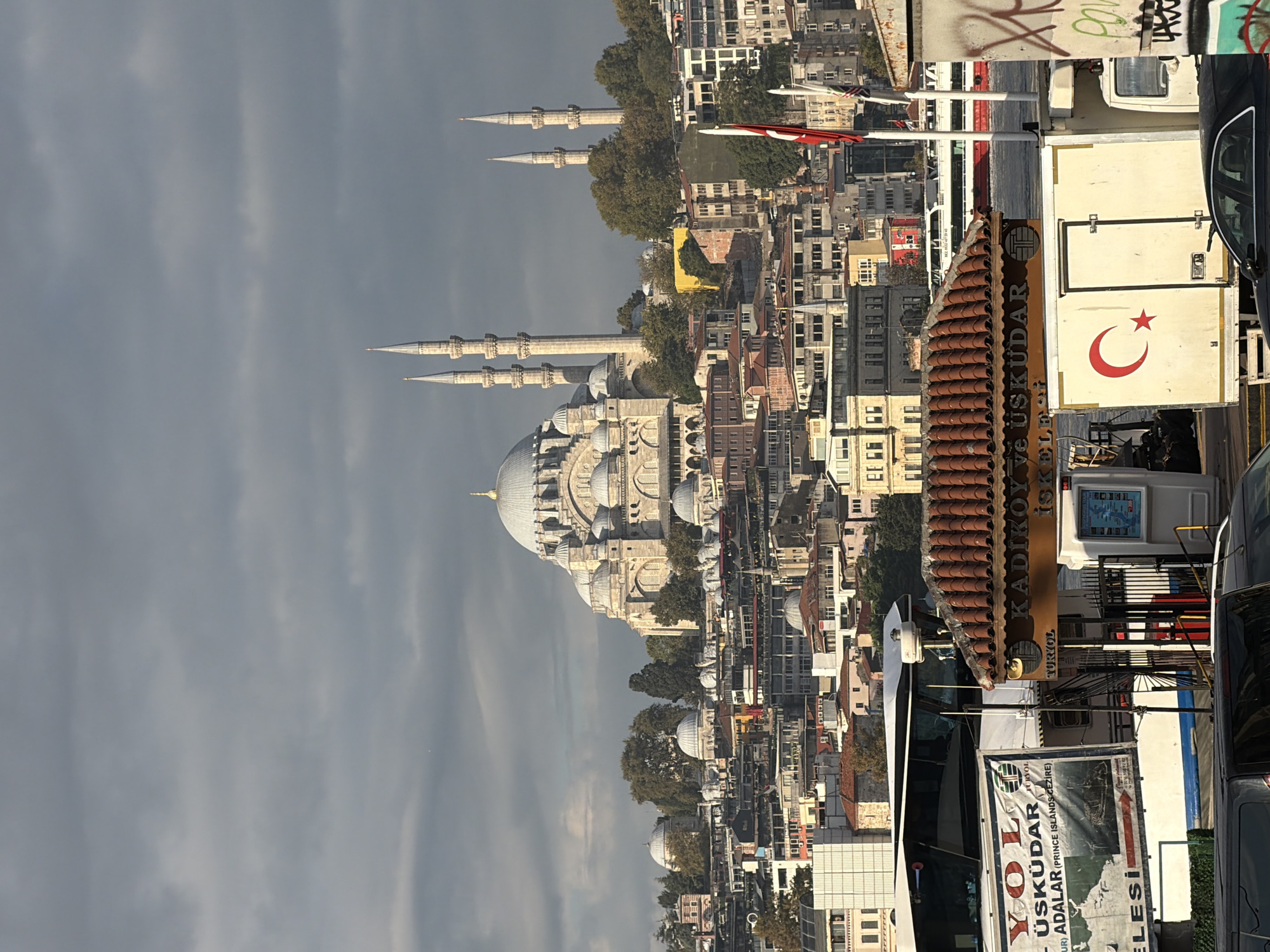A panoramic view of the cityscape of Istanbul, Turkey, with a prominent mosque dominating the skyline. The mosque features a large central dome and multiple minarets, characteristic of Ottoman architecture. The surrounding area is densely populated with buildings of varying architectural styles and colors, indicative of urban sprawl. In the foreground, a ferry terminal is visible, with signs indicating destinations such as "KADIKOY ve ÜSKÜDAR" and "YOL ÜSKÜDAR ADALAR". A white truck with a Turkish flag symbol is parked near the terminal. The sky is overcast with dramatic clouds, suggesting either an approaching storm or a recent one. The overall impression is a bustling urban environment with a strong historical and cultural presence, as evidenced by the mosque and ferry services. The specific location of Beyoğlu, Turkey is mentioned, but this view is more representative of the Sultanahmet or Üsküdar districts, known for such prominent mosques and ferry terminals.